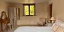 A cosy bedroom featuring a large bed with decorative pillows, a wooden vanity with a mirror, an armchair, and a window with light curtains. Soft, warm