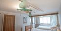A bright, clean hospital room with two neatly made beds, ceiling lift system, wooden door, framed artwork, and curtained window.