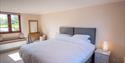 Bright bedroom with a large bed, grey headboard, and white bedding. A lit lamp on a bedside table adds warmth. A window bench and mirror complete the