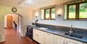 Bright kitchen with white cabinets, blue countertop, and black stove. Sunlight streams through two windows. Archway leads to a hallway. Rustic, cosy f