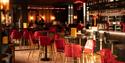 Warmly lit bar with red velvet chairs and round tables. A modern, inviting ambiance is created by hanging lights and a stocked bar with glassware.