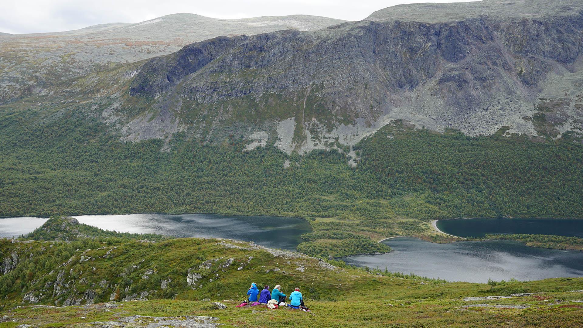 Four persons sit in the grass and enjoy a wild landscape with lakes and steep mountain sides.