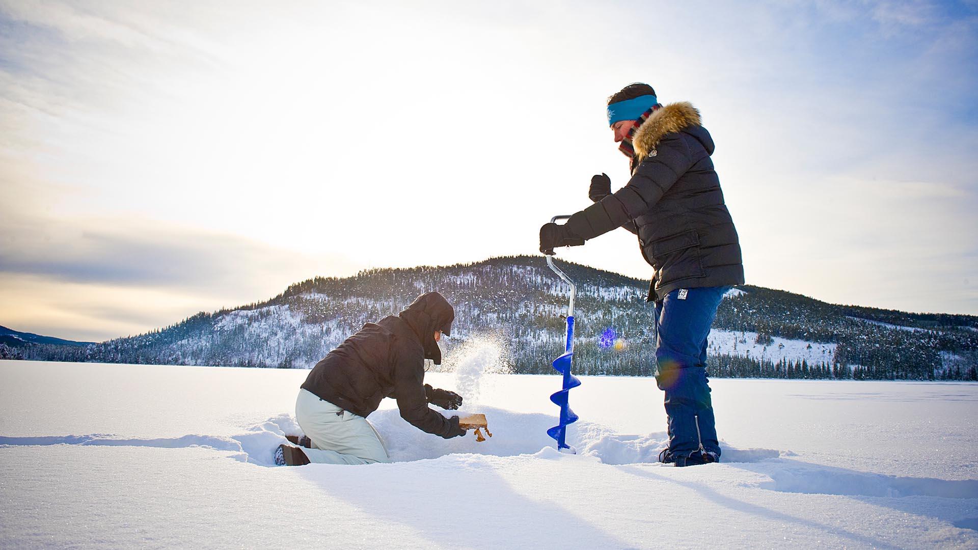 To isfiskere på snødekt vann med fjell i bakgrunnen.