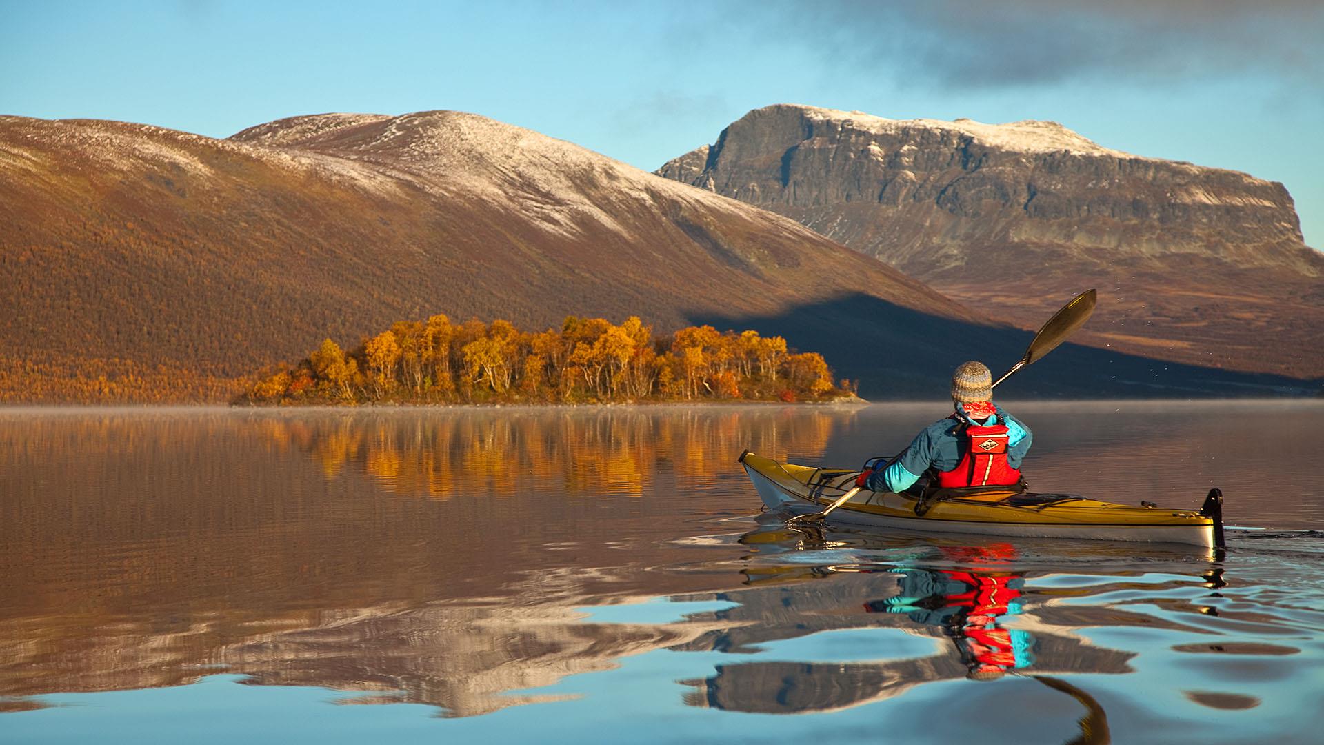 Eine Person im Kajak paddelt an einem Herbstmorgen auf einem spiegelblanken Bergsee auf eine Insel zu, die mit orangegefärbten Birken bewachsen ist. Im Hintergrund erheben sich mächtige Berge mit steilen Abhängen und dem ersten Neuschnee auf den Gipfeln.