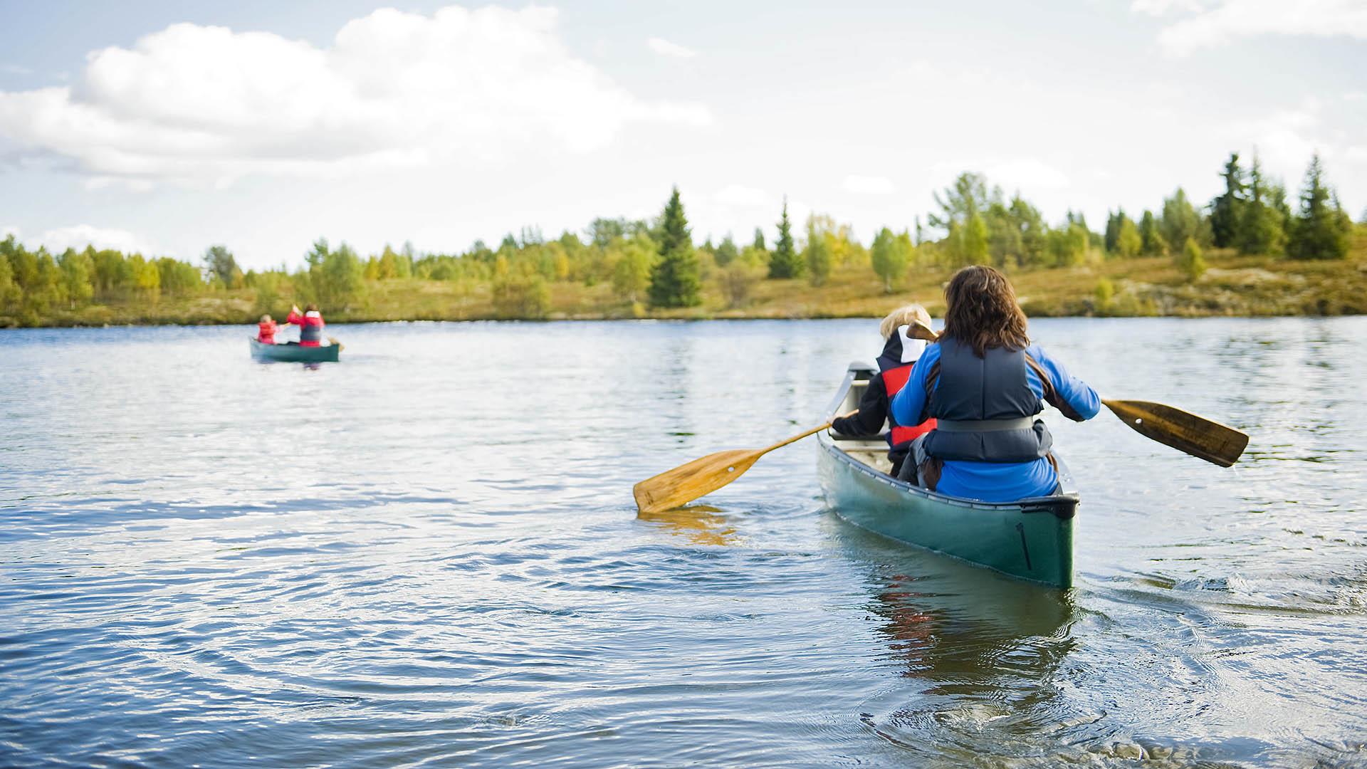 Two canoes, one close by and one further away, on a lake with åpen birch and spruce forest in the background.