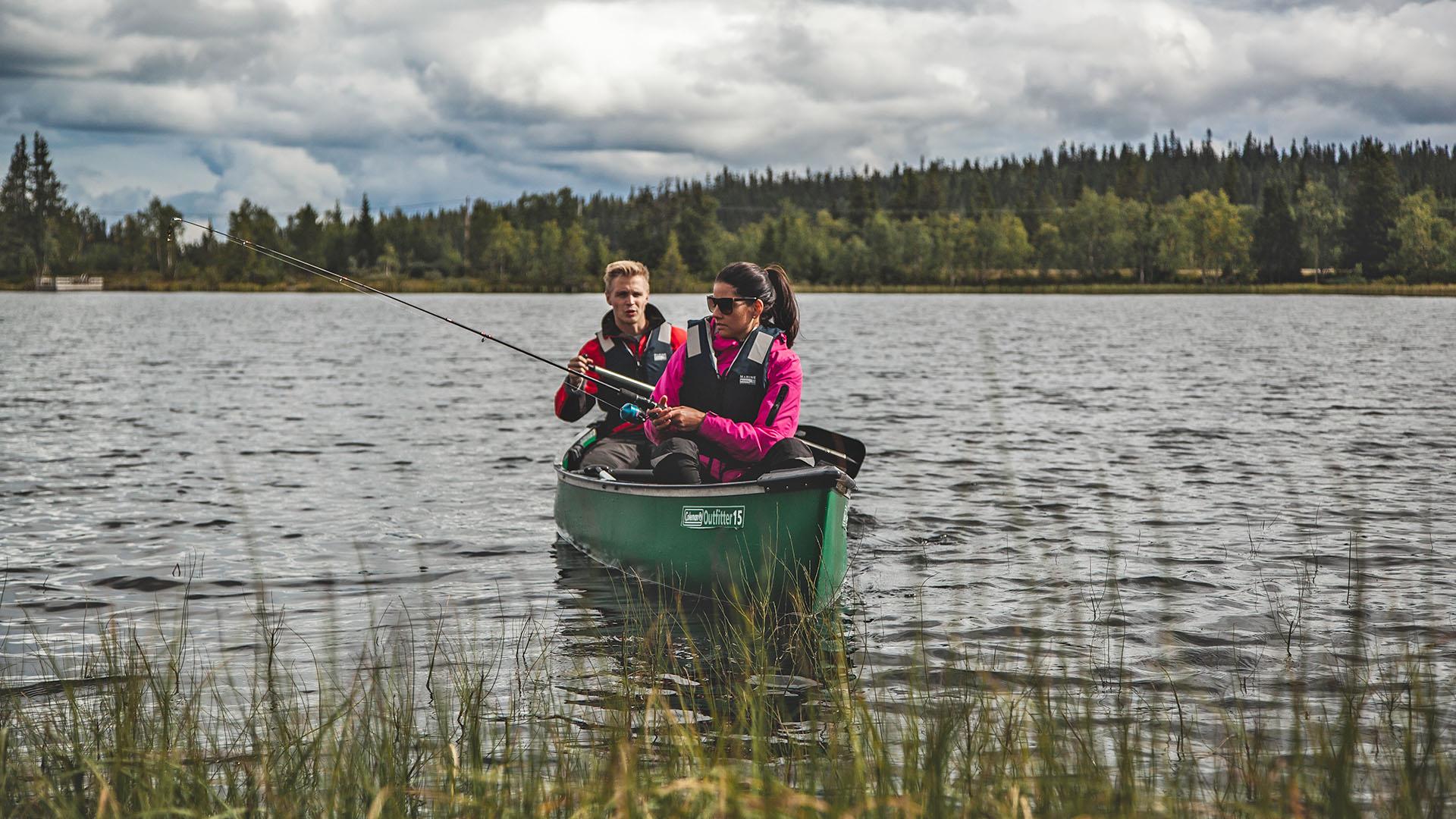 Two persons in a canoe on a lake close to shore with some vegetation in the front. One person fishes with rod, the other paddles.