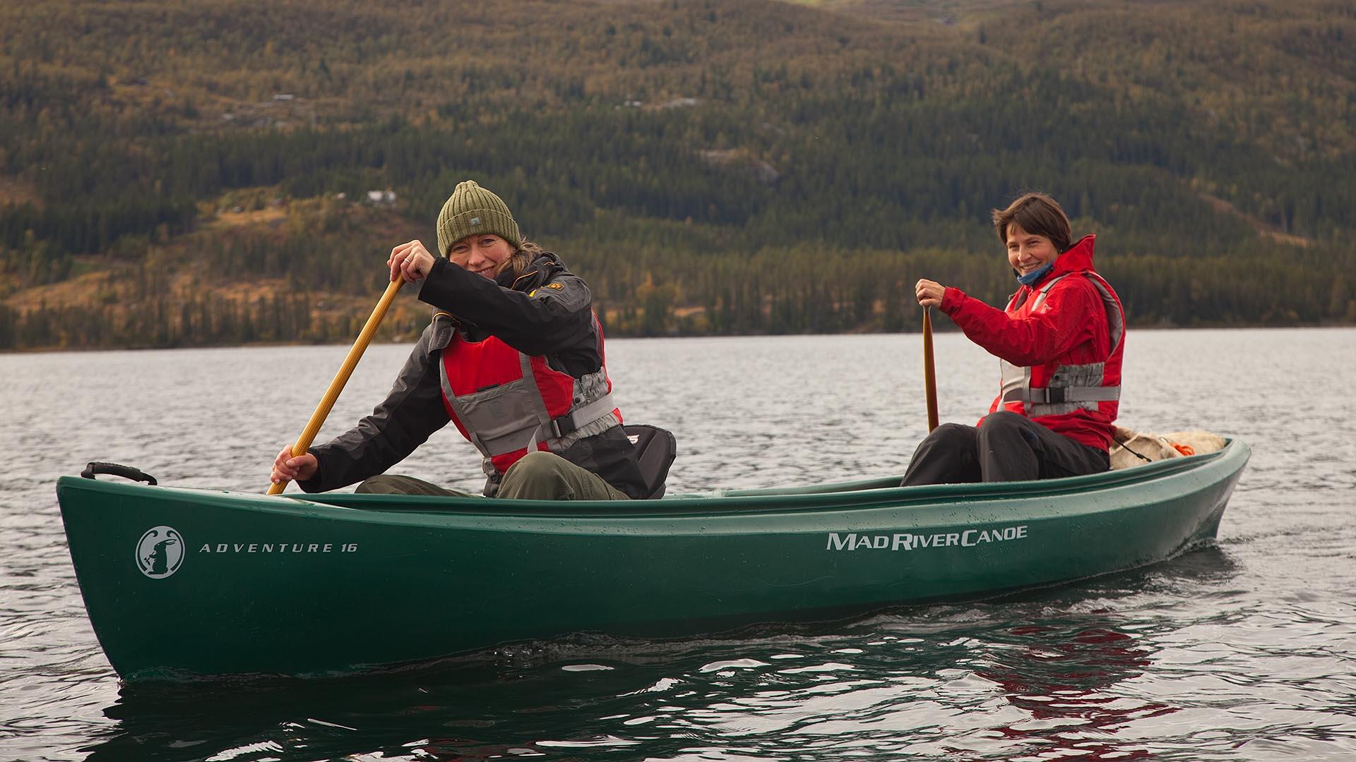 A green canoe with two persons inside, facing the photographer, on a lake in autumn.