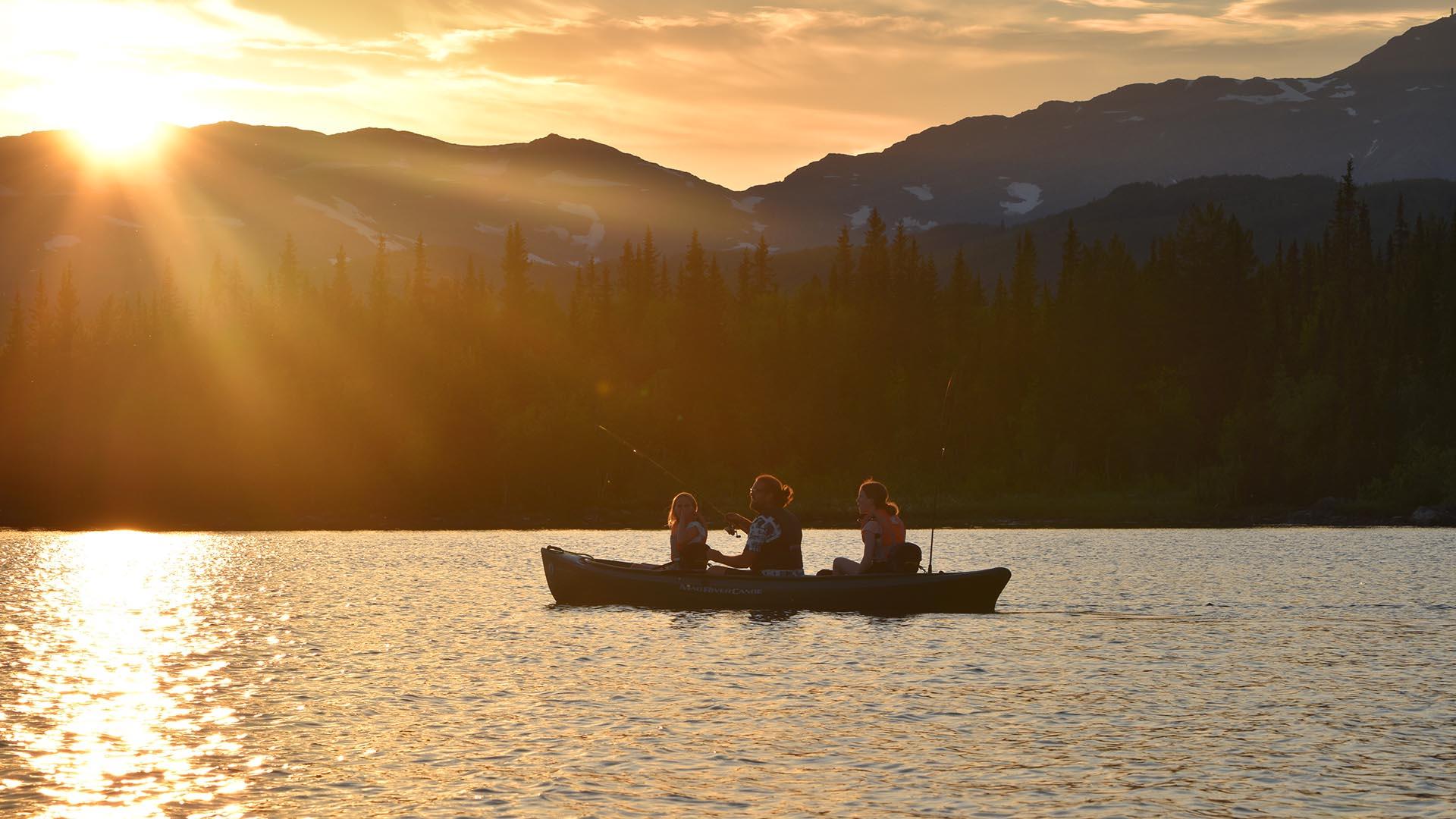 A canoe with three persons that fish with rod on a lake with mountains in teh background. The setting sun paints the sky and the water in yellow and orange.