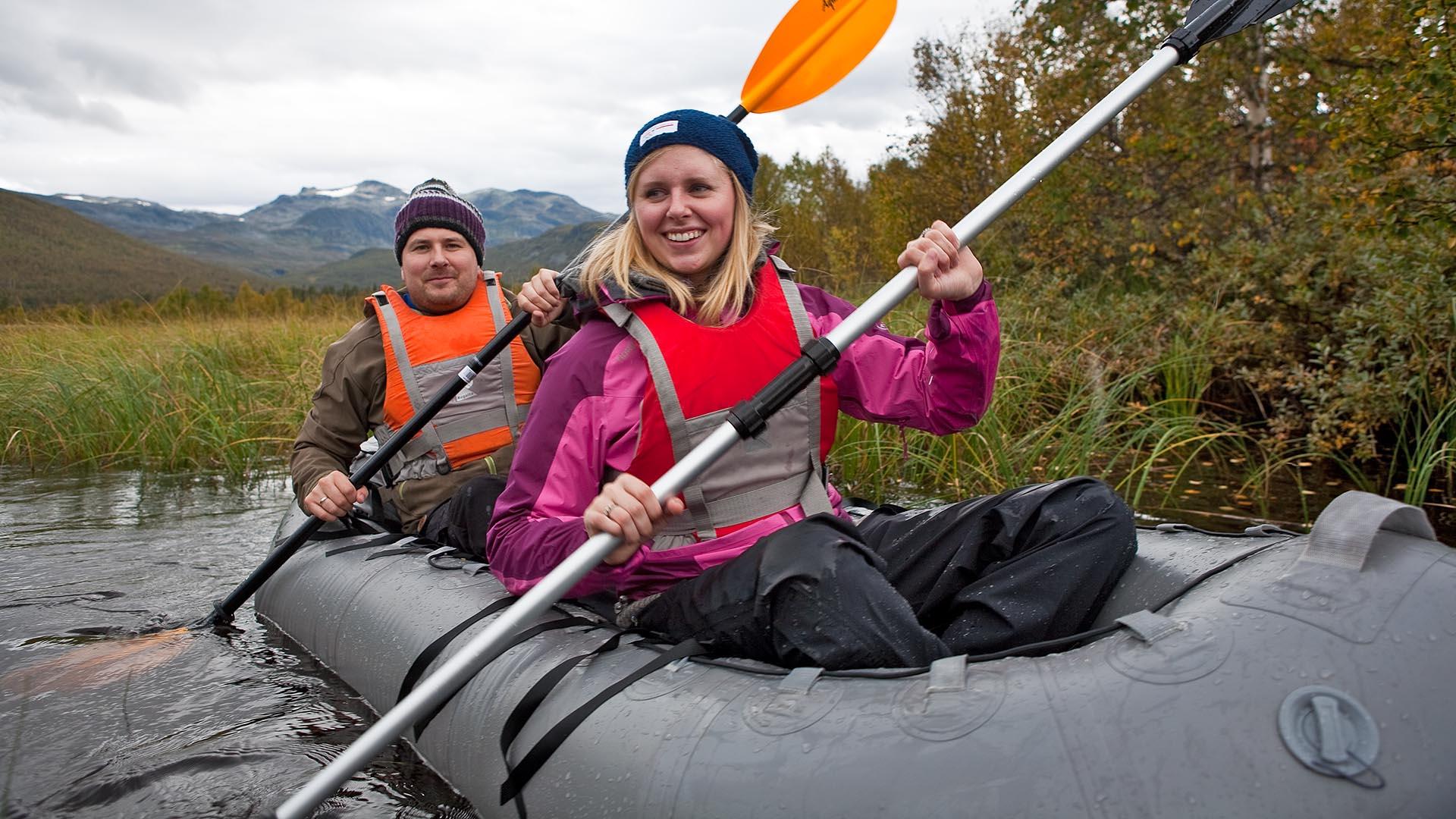 Nærbilde av to personer i en packraft på en liten elv med siv og busker langs breddene. Fjell i bakgrunnen i det fjerne.