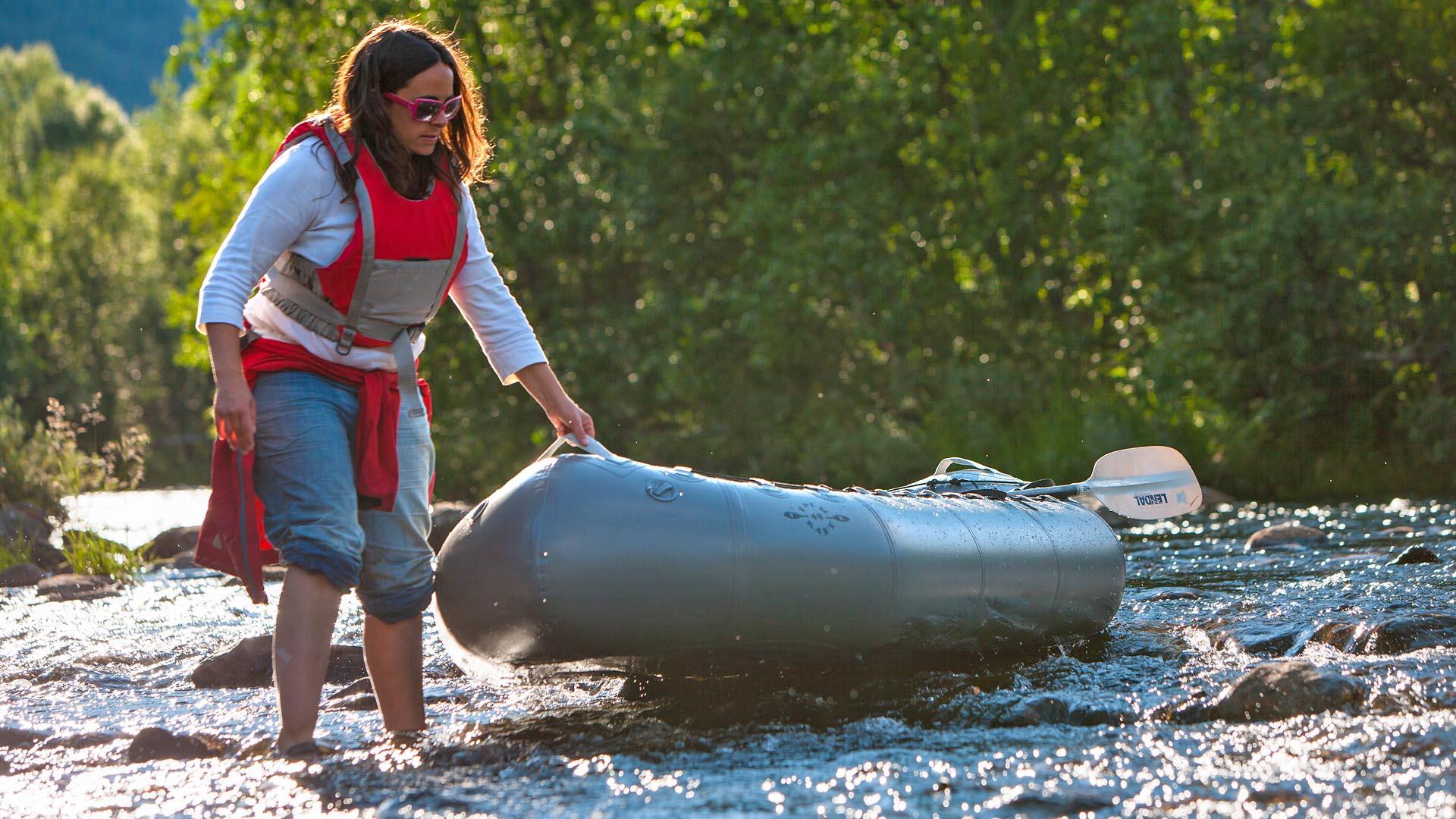 En dame med rød redningdvest vasser og drar en packrafting flåte over en passasje med lav vann og steiner i elva.