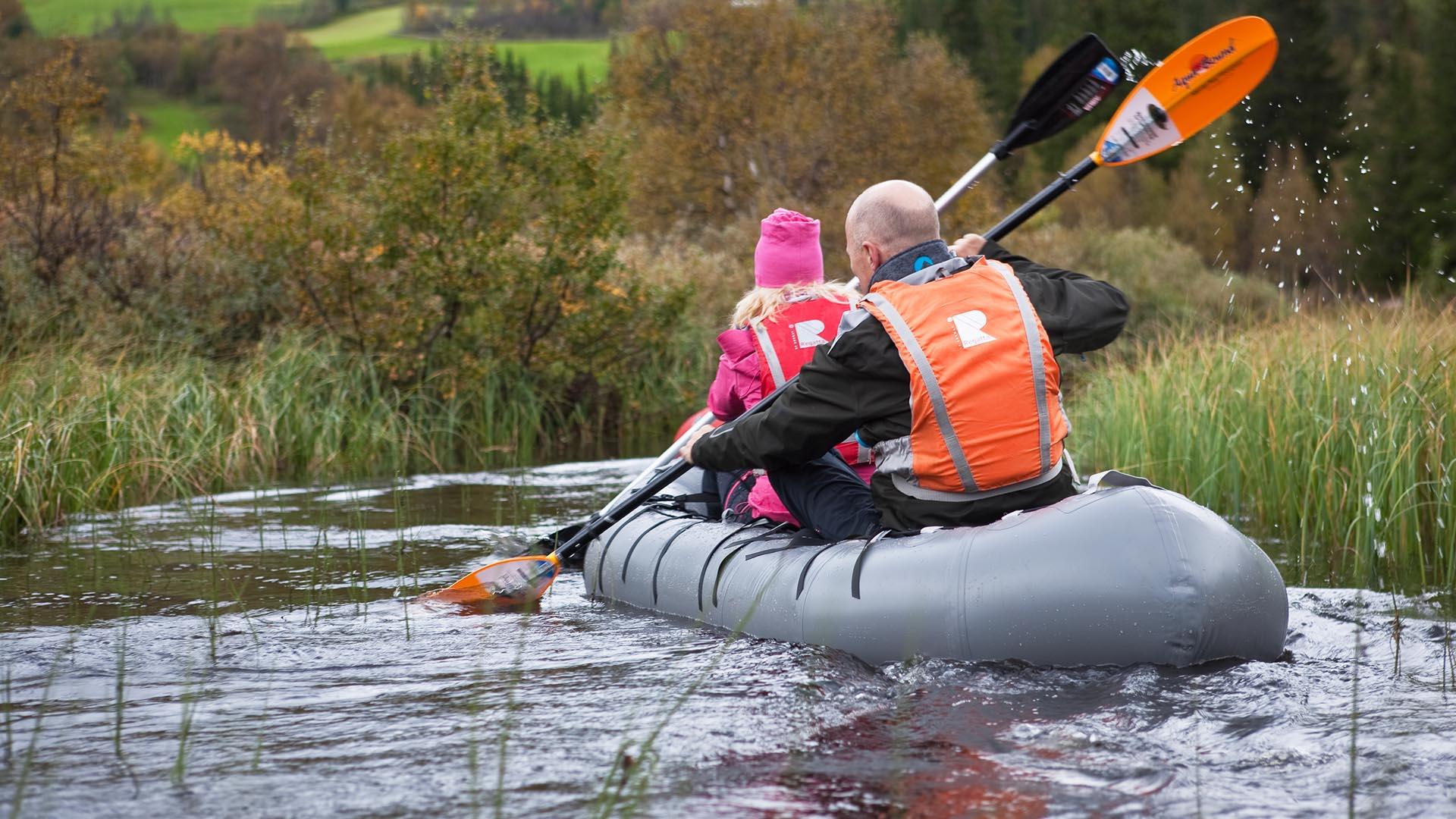 Zwei Personen in einem Packraft auf einem kleinen Fluss, dessen Ufer mit Schilf und Büschen bewachsen ist.