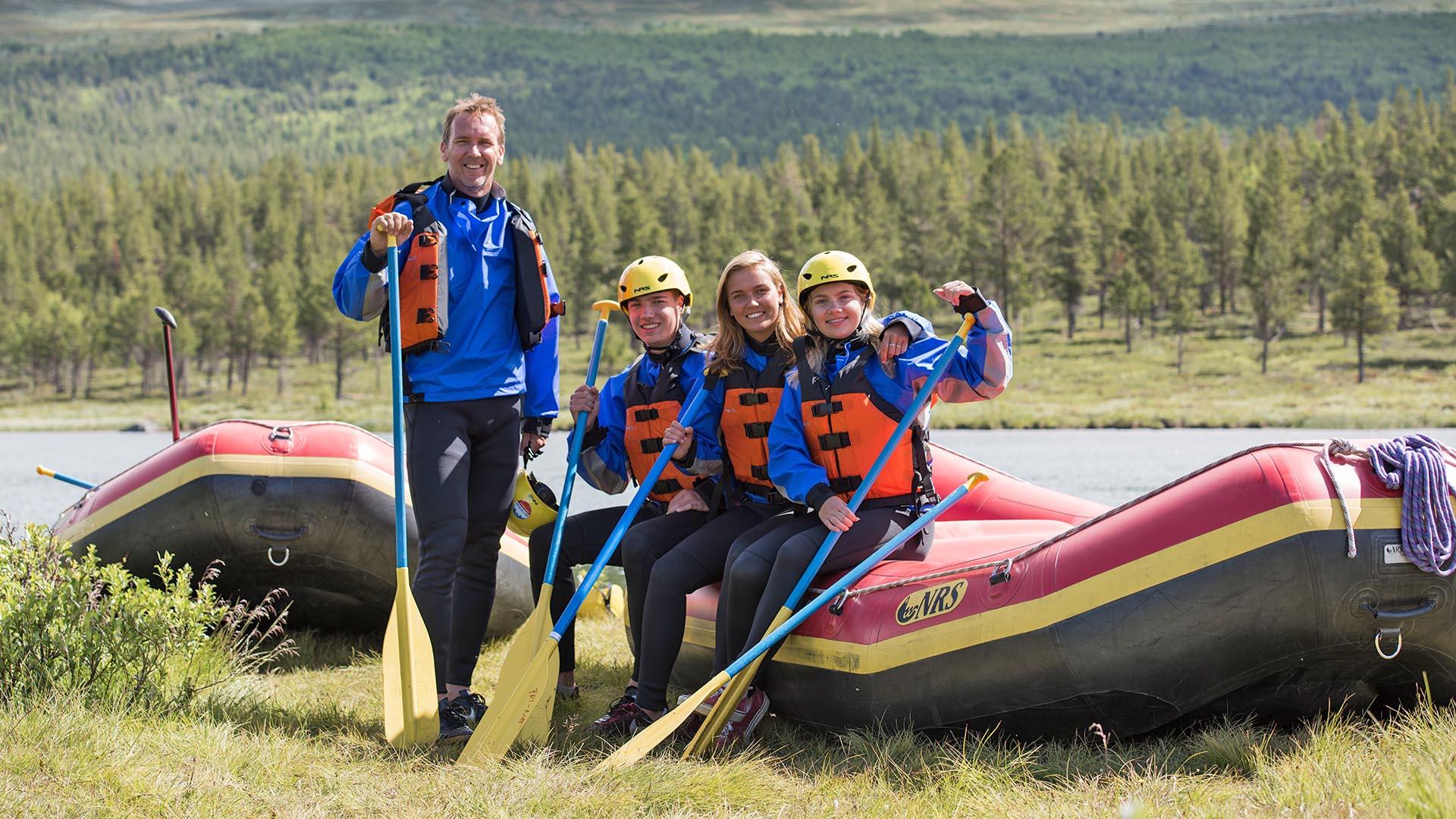 Et team av fire raftingguider poserer ved to flåter på en elvebredd.