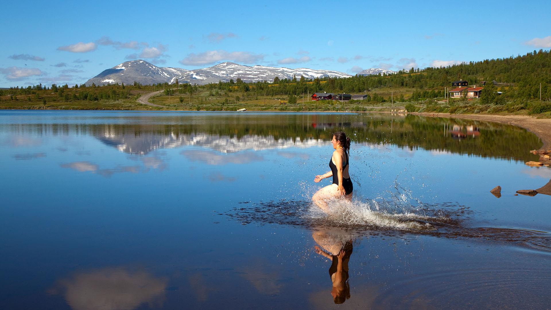 En dame i badedrakt går ut i et speilblankt fjellvann med sandstrand. Noen stølshytter og fjell i bakgrunnen.