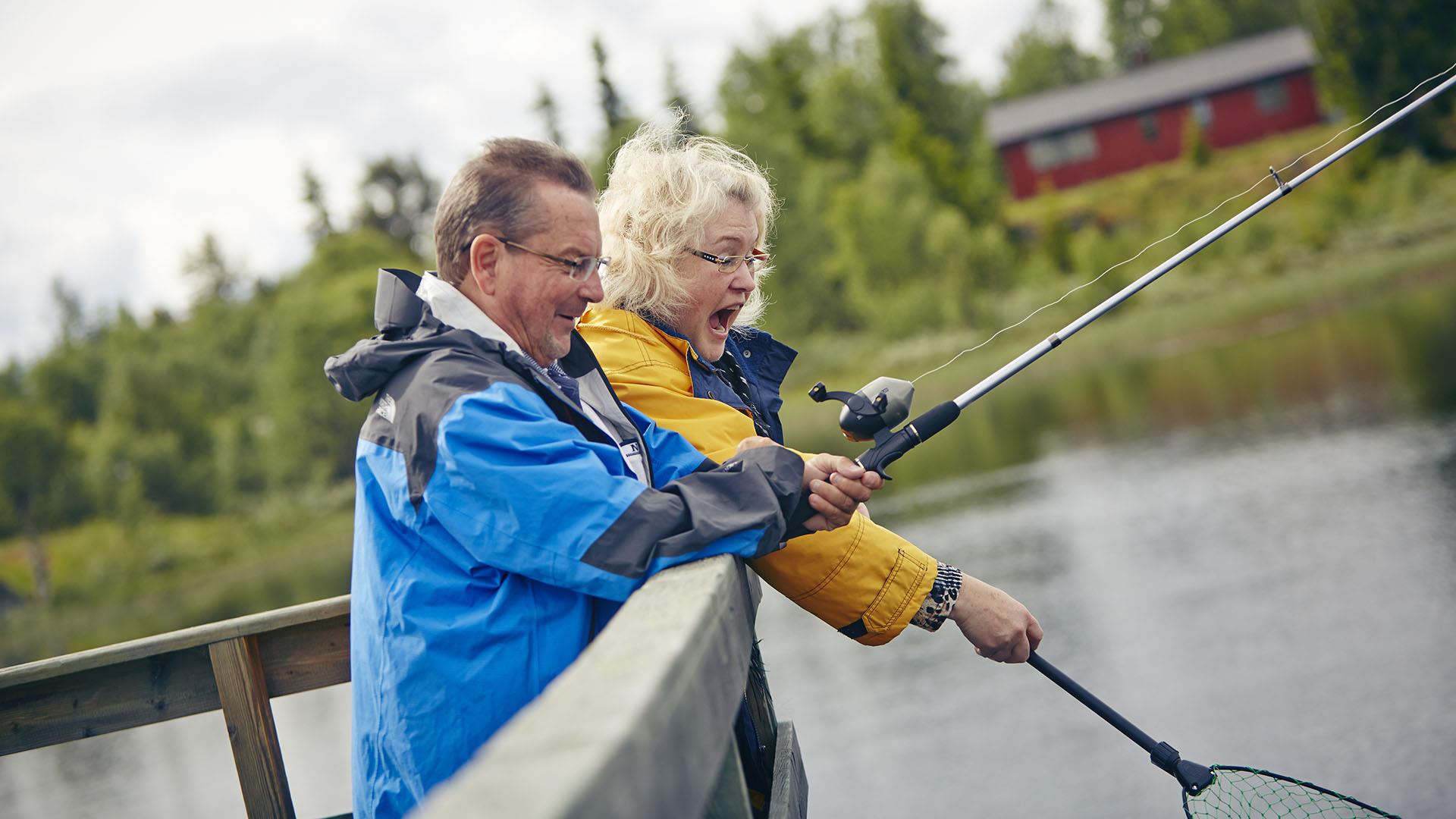 En voksen dame og mann står på en fiskebrygge med fiskestang og hov, full av begeistrng mens drar iland fangsten.