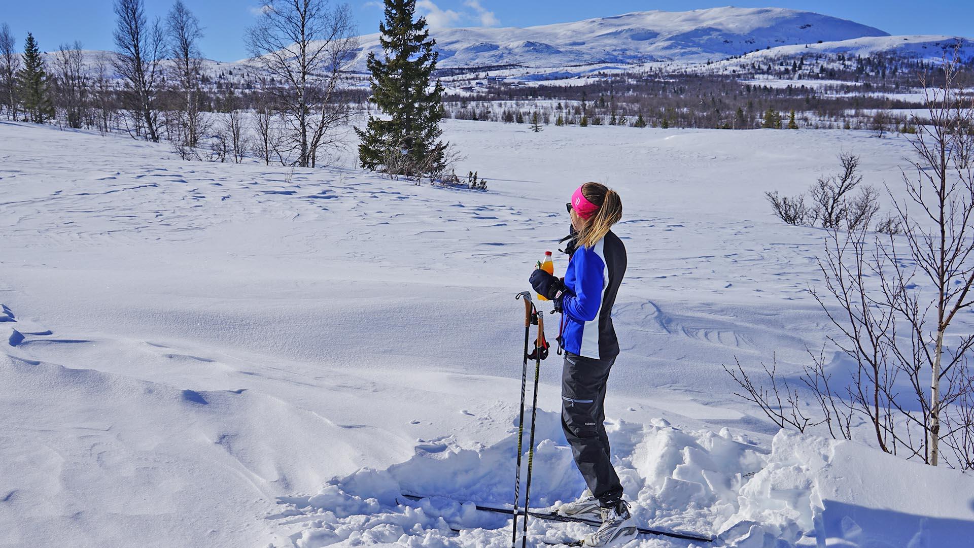 Kvinne tar seg en velfortjent rast langs skiløypa, med skog og fjell i bakgrunnen.