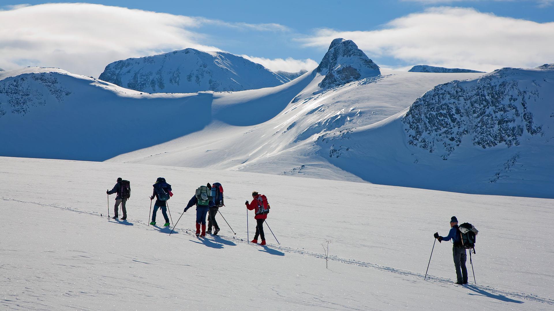 En gruppe fjellskigåere med sekker langs en kviteløype i Jotunheimen foran breer og høye, spisse fjell.