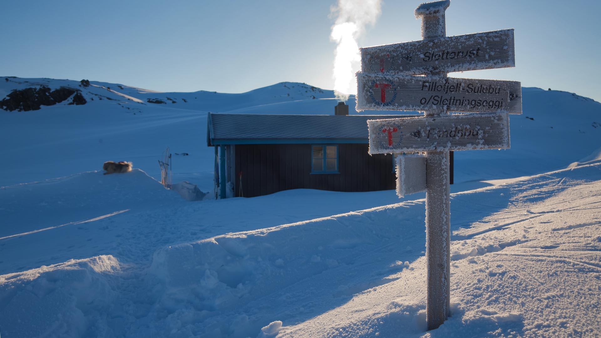 En ensom hytte i vinterfjellet med røyk fra pipa og en nedrimet skiltpåle. Lav vintersol gir mye skygge.