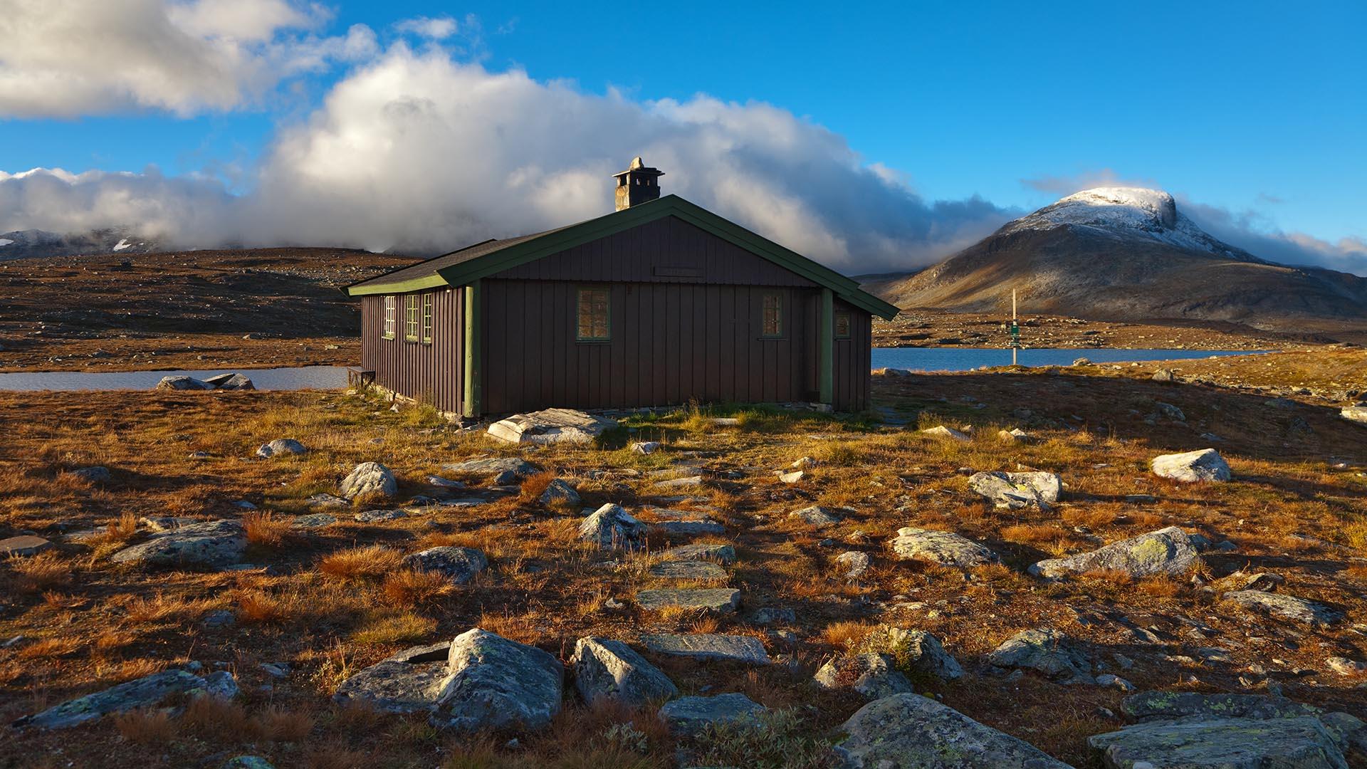 En brunmalt hytte stor i fjellet ved et vann, men et snødekt fjell i bakgrunnen.