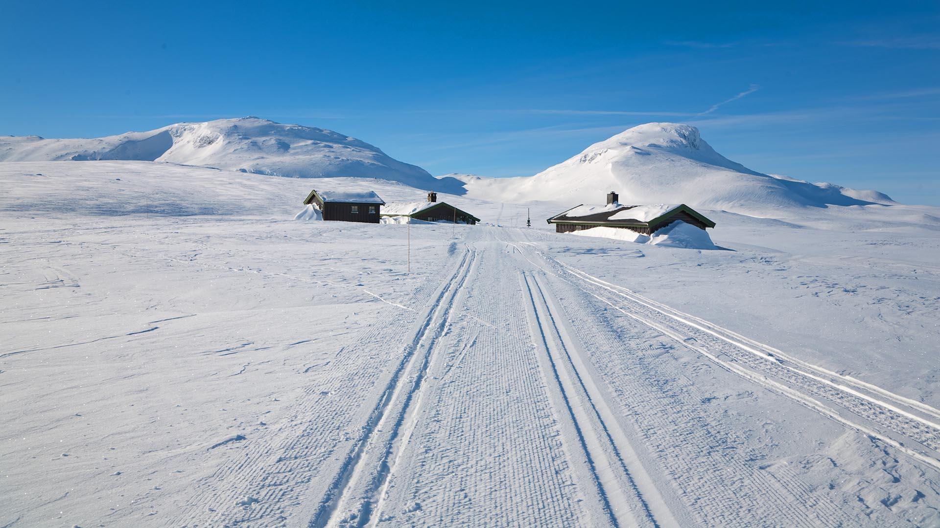 En skiløype leder rett mot en hytte i fjellet. To fjelltopper ligger bakenfor.