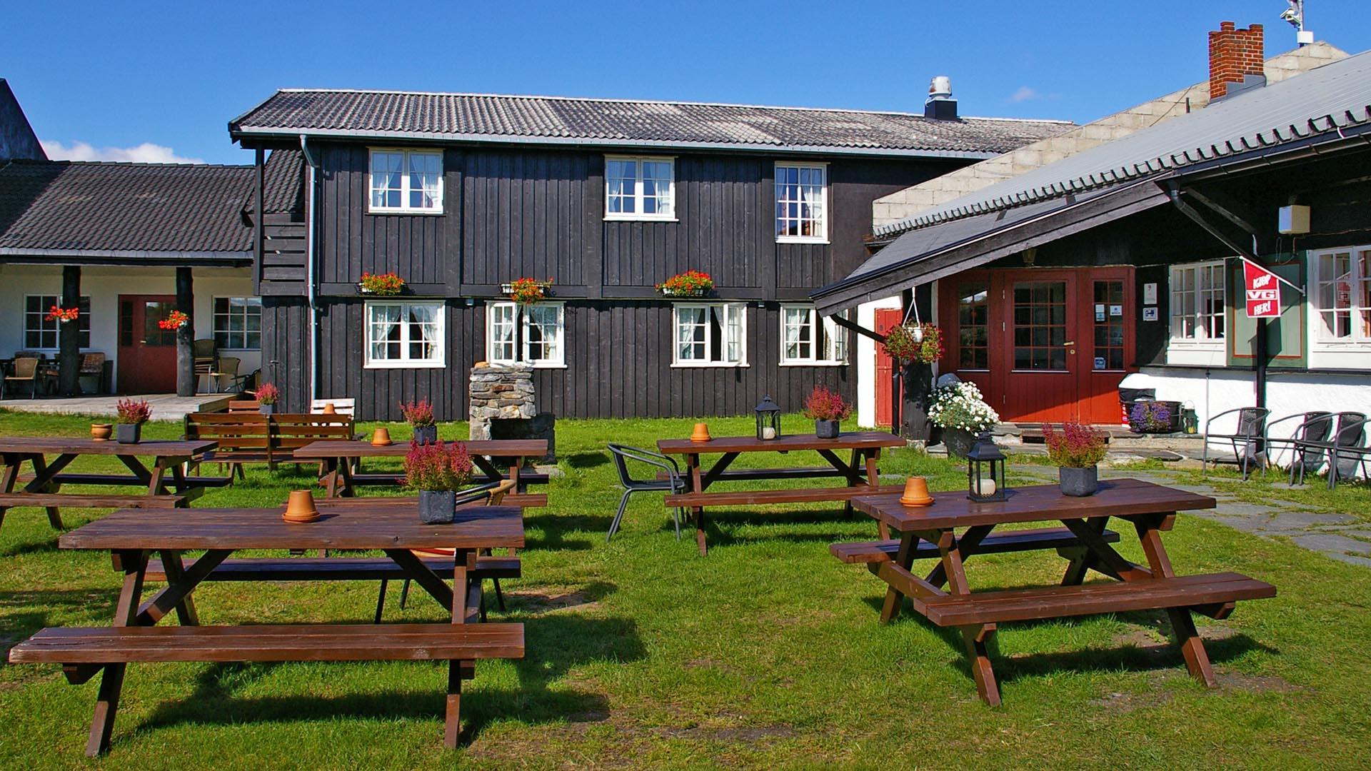 Gressbevokst gårdsplass bak en fjellstue med flere benkebord.