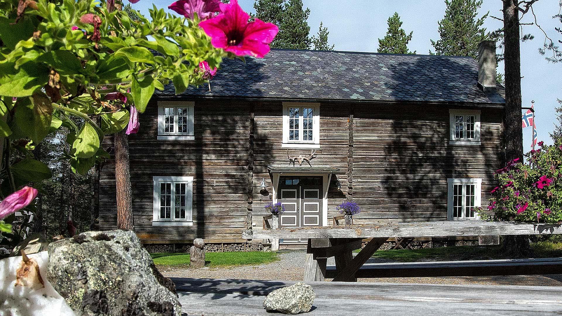 Laftet gårdsbygning med hvite vinduskarmer og skifertak på et tun med blomster i hengepotte