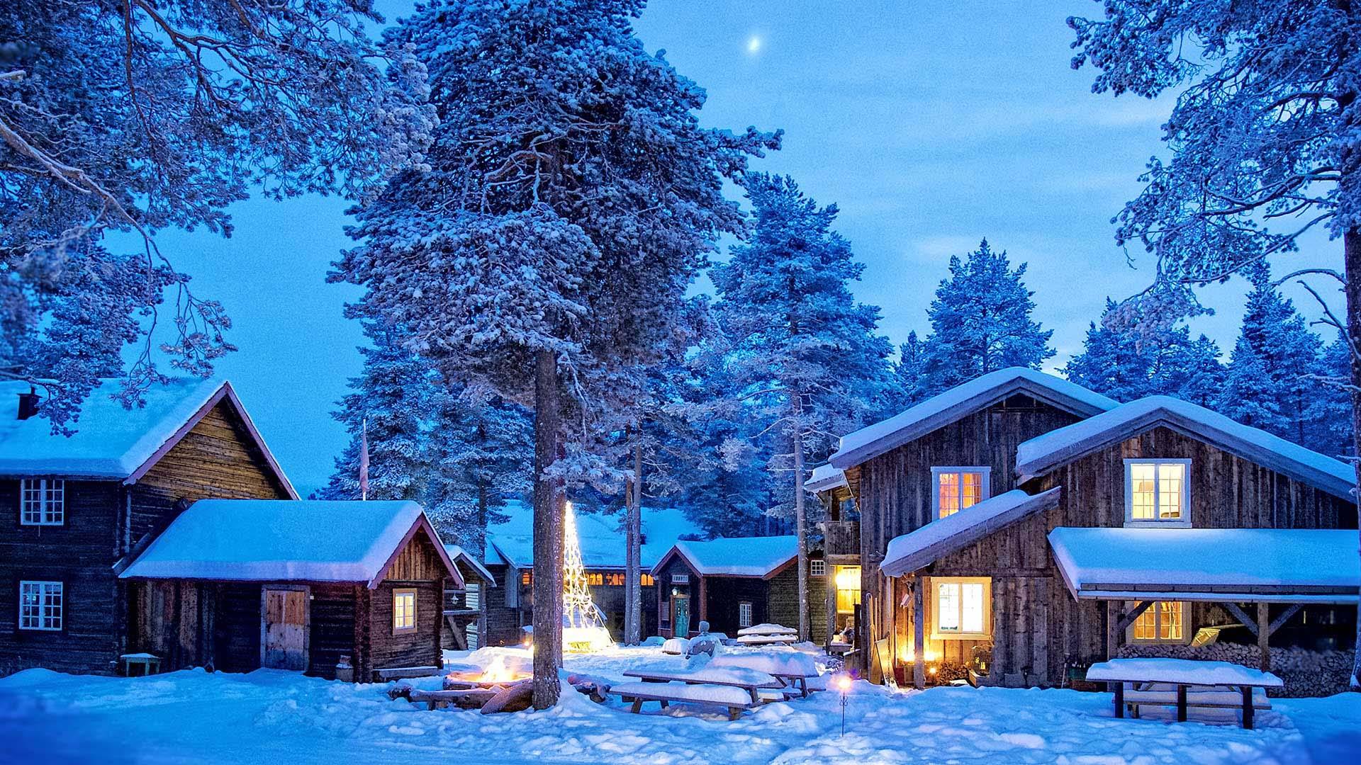 Opplyst tun av laftede bygg som er et hotell i åpen furuskog en vinternatt med snø og fullmåne