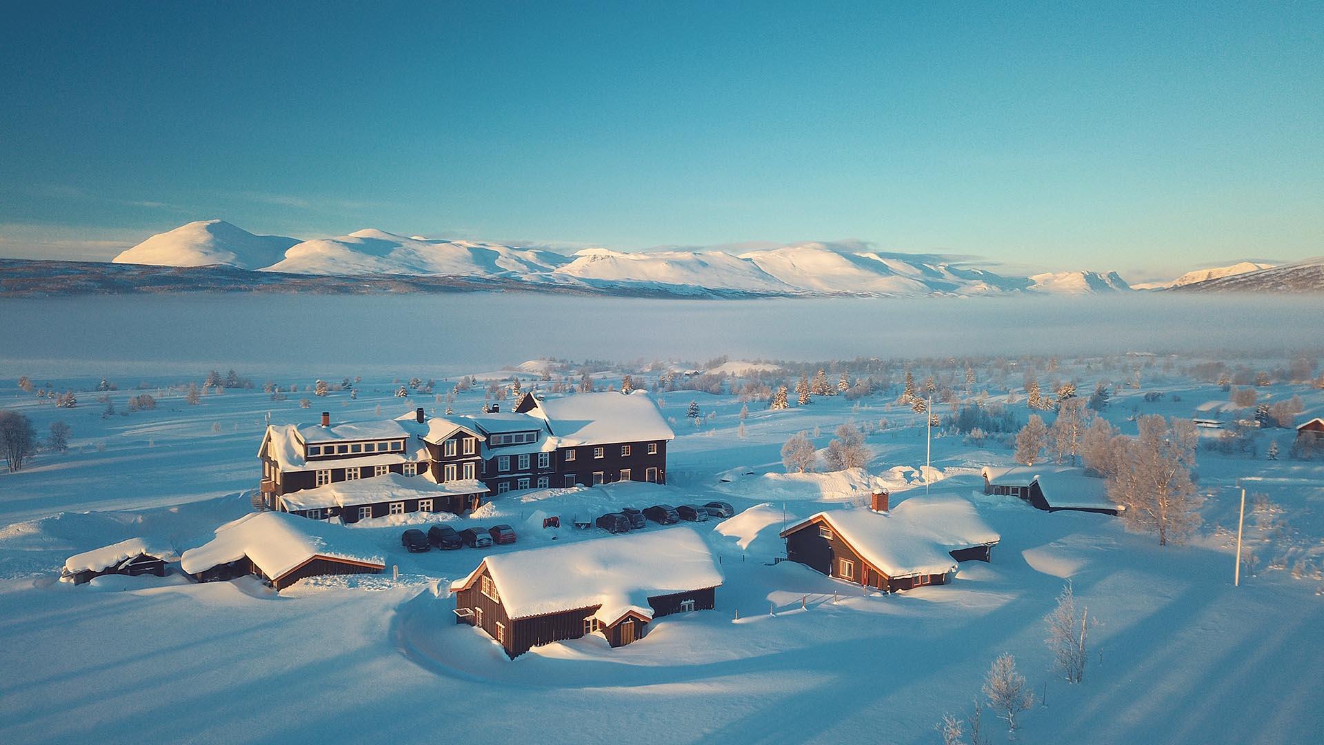 Luftfoto von einem Hotel mit umliegenden Hütten in verschneiter Fjellandschaft und weichem Licht mit schneebedecktem See und Bergen im Hintergrund