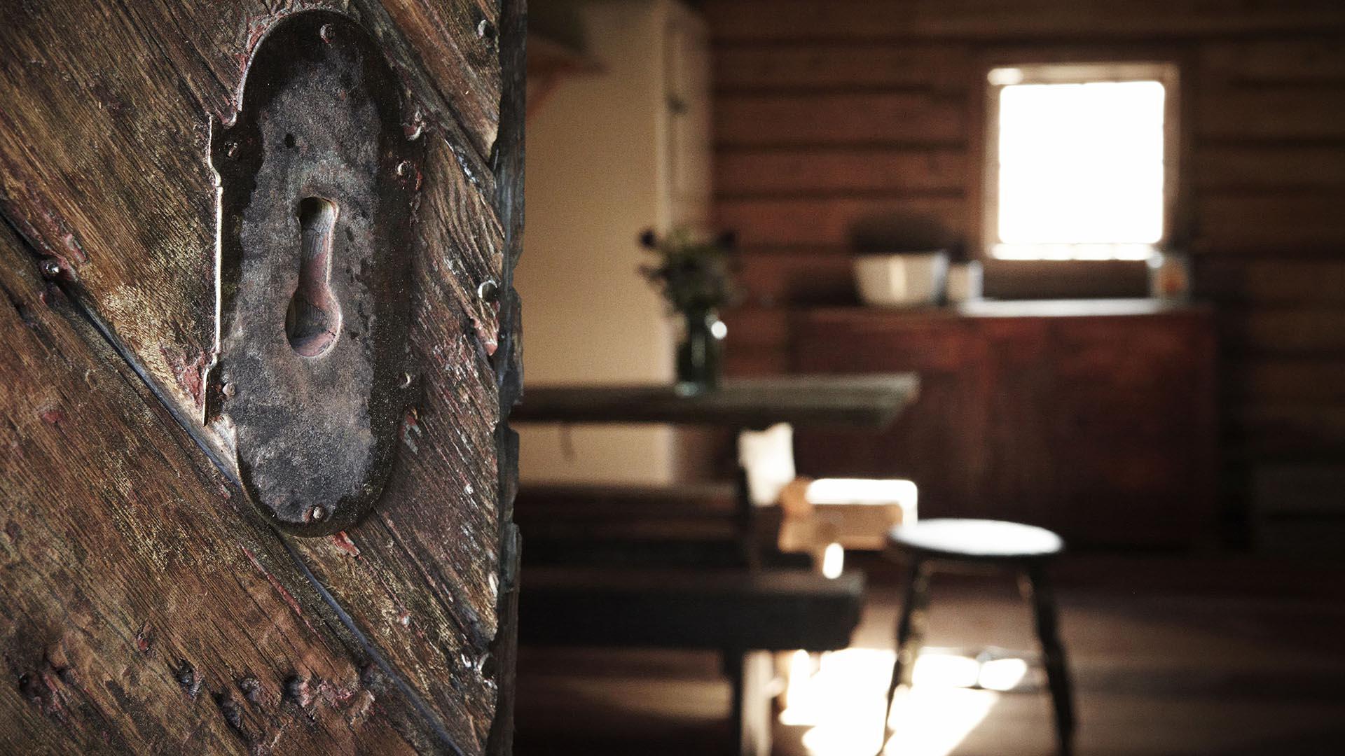 Detail of a iron mounting on an old wooden door which is half open and grants a blurry view into the room with old furniture lit up by the sun through the window.