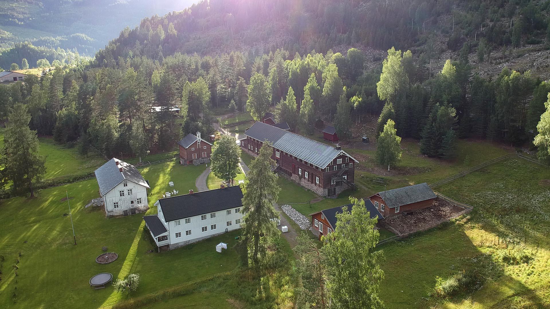 Luftfoto av en storgard med hovedhus, kårbolig og stor låve. Sola skinner gjennom tretopper på en grønn plen.