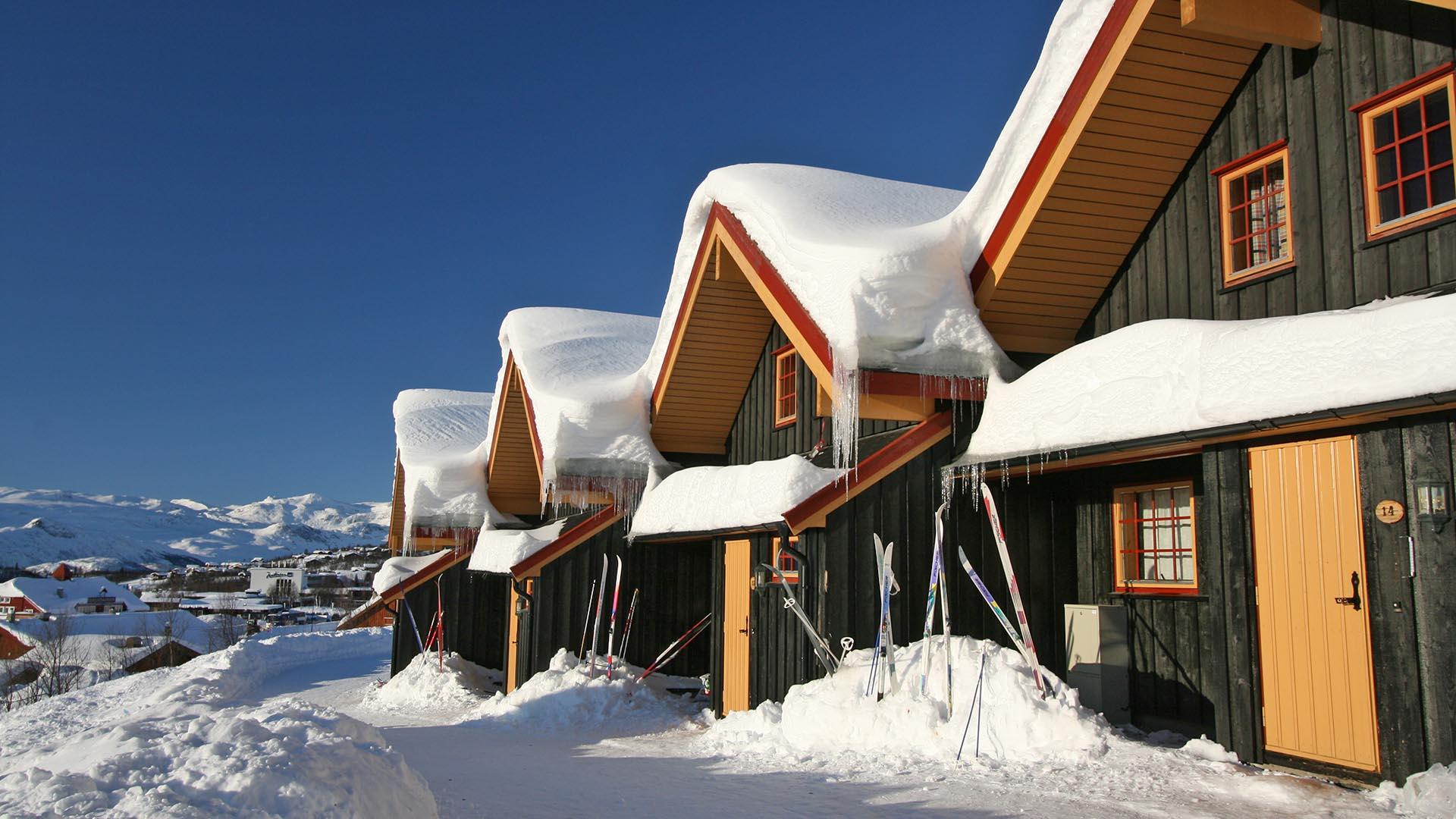 Dark brown wooden attached cabins with lots of snow on the roofs on the sunny side of the hill of a mountain village (seen in the background).