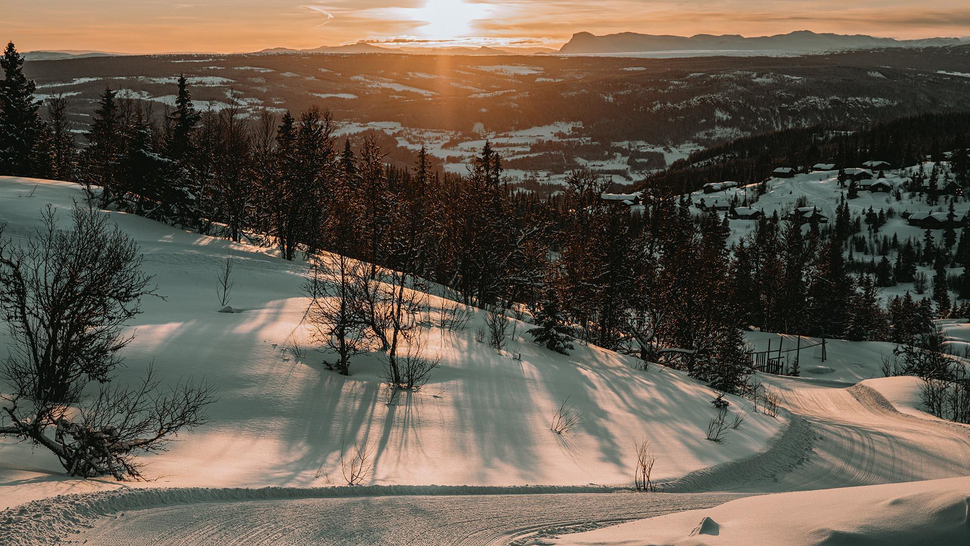 View from a hill over a valley towards the sun which is setting behind mountains on the horizon. The snow on the ground is painted yellow in the evening light.