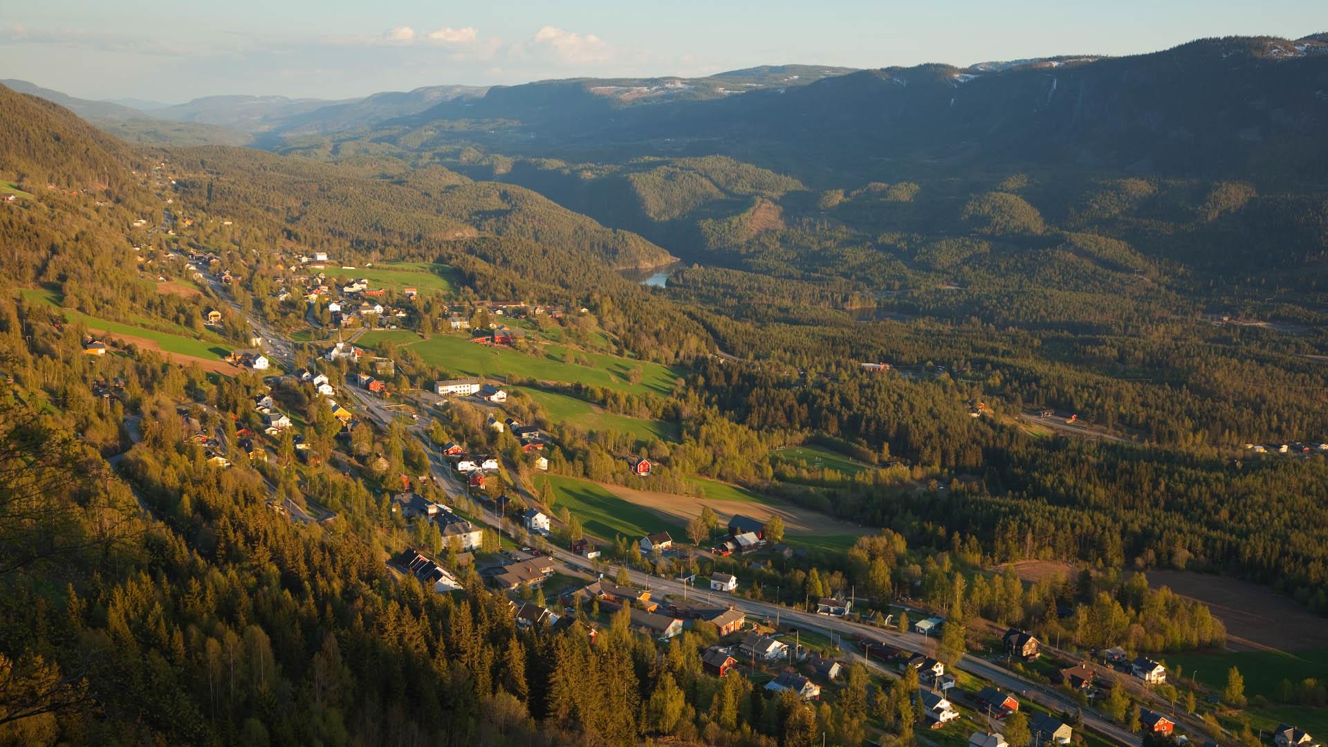 View over a valley with forested hills and a village with green fields that stretches alongside a road