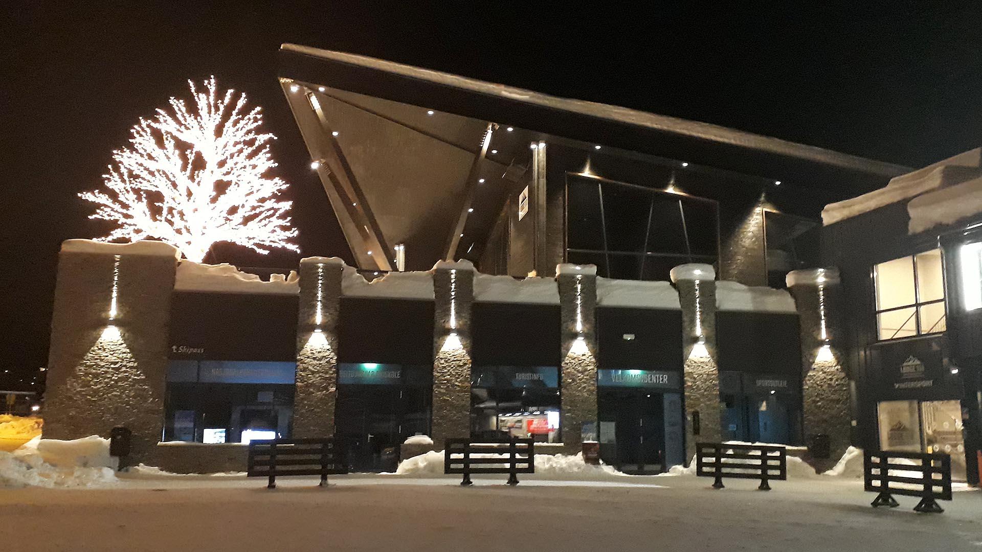 A modern building in a black winter night that is lit up by a large tree-shaped light-sculpture on the roof terrace and several lights that seem to flow down on the facade like waterfalls