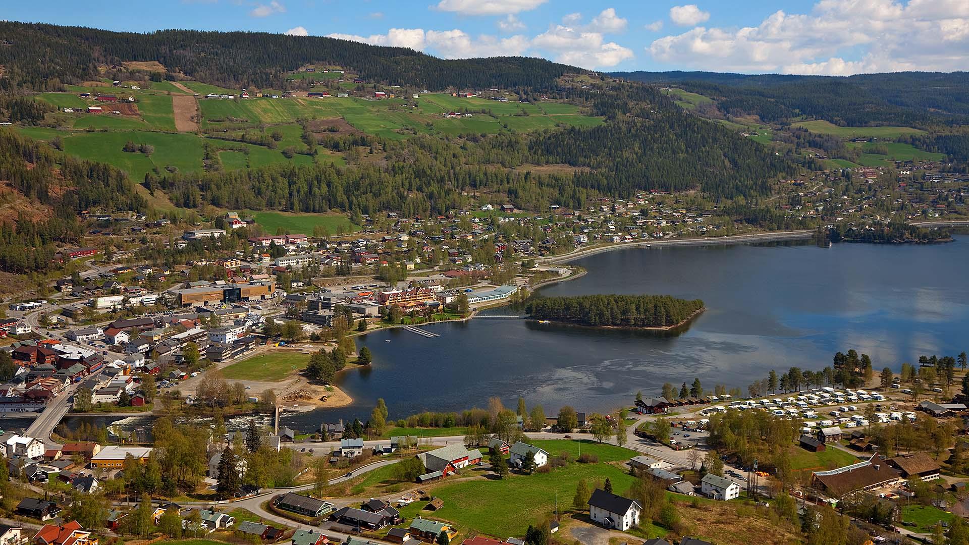 Utsikt utover en liten by som ligger ved utløpet av en elv i et stort vann, med en øy i vannet og grønne jorder i liene bakenfør