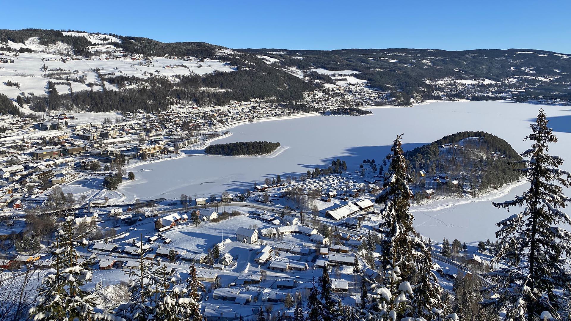 Utsikt fra en høyde over en liten by ved et vann, men en halvøy og en øy ute i vannet. Landskapet og vannet er snødekt og himmelen blå. Noen høye grantopper i forgrunnen.