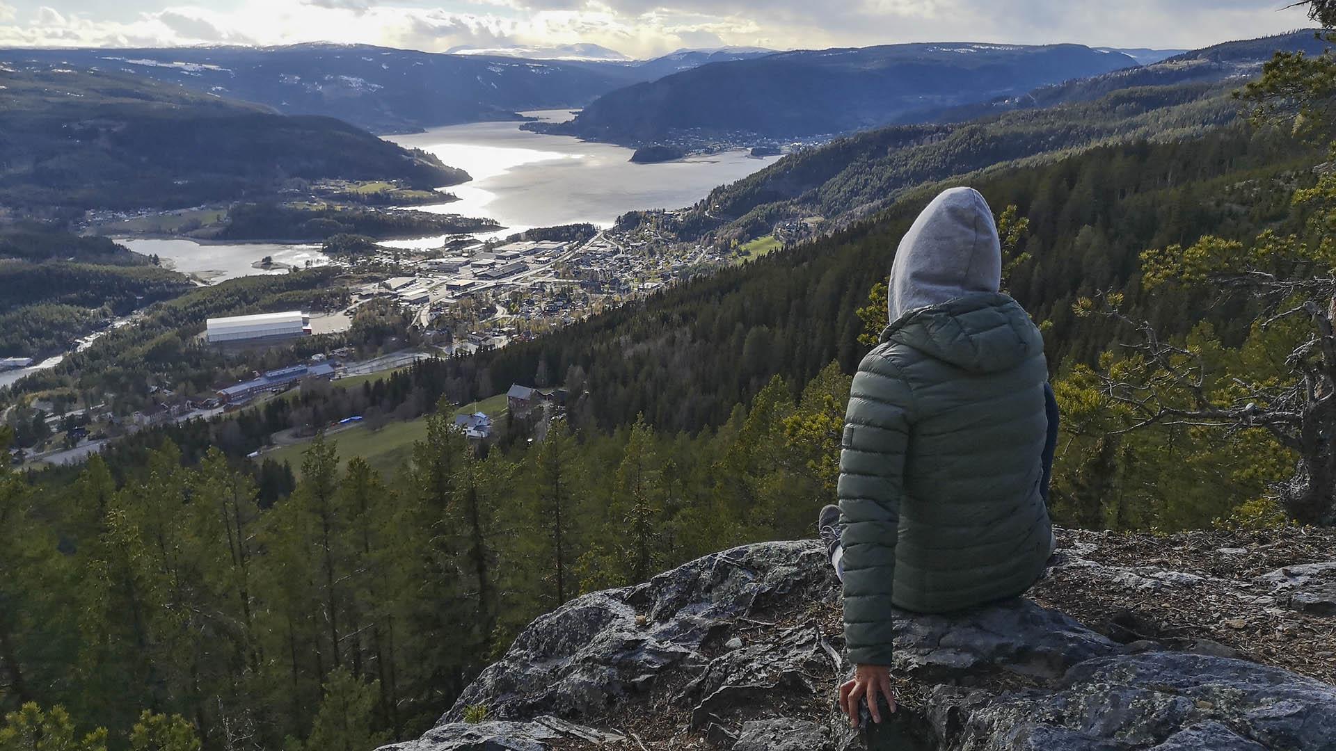 En ung jente med hettegenser sitter med ryggen til på en fjellknaus ovenfor en skogkledd dalside og skuer oppover dalen med et tettsted og et stort vann mellom dalsidene bakenfor. Motstående sol glinser i vannet.