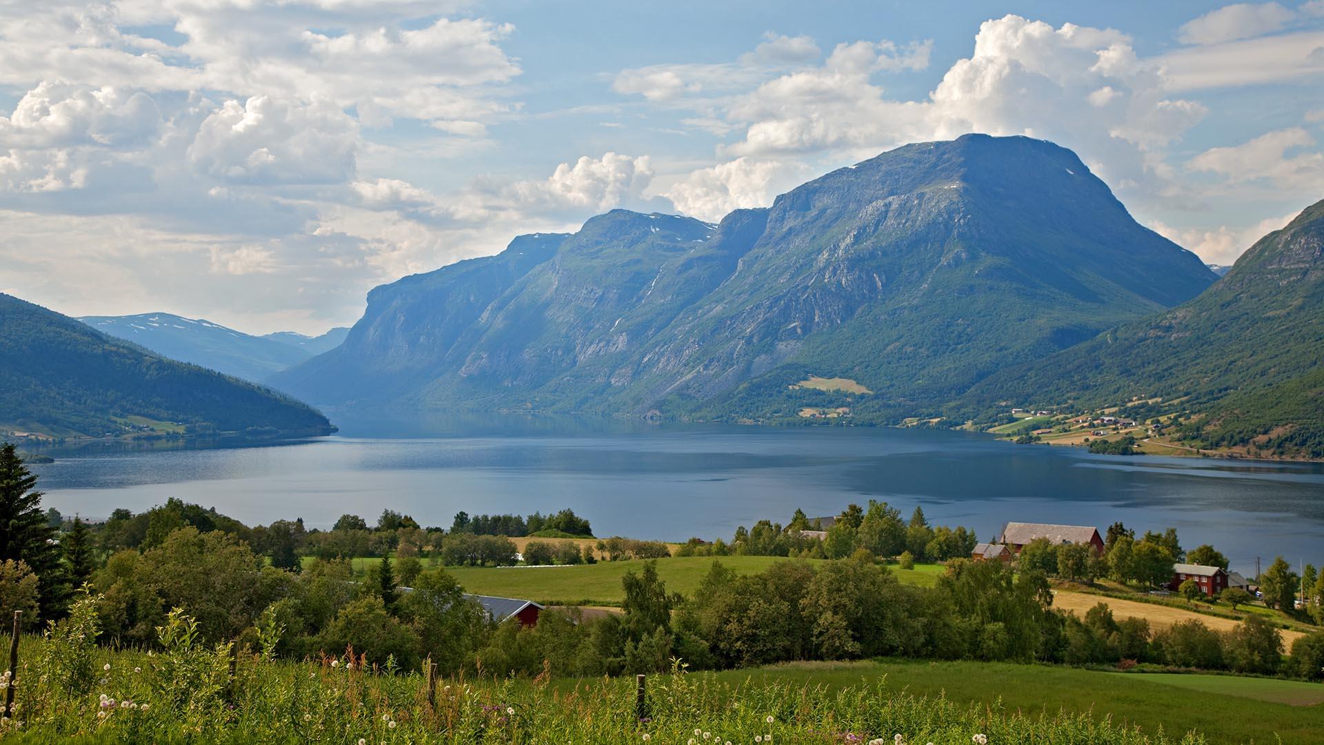 Kulturlandskap med grønne enger og jorder med noen trær foran et stort vann med et massivt fjell bakenfor vannet.