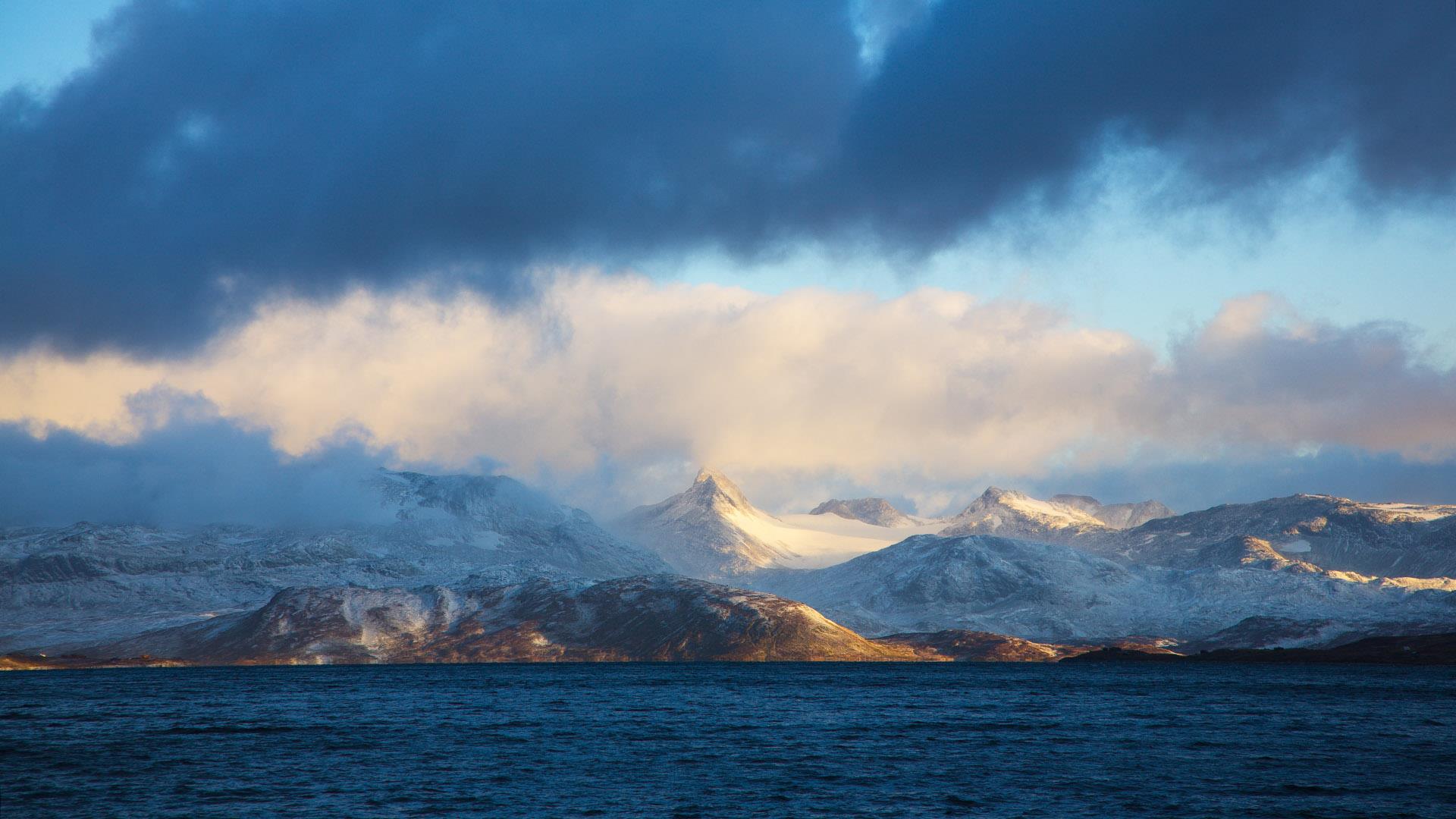 Late autumn in the high mountains. A large lake in the forground with dark blue water and behind high pointed mountains with autumn-coloured bases and snow-covered summits. White and dark grey clouds and some blue sky inbetween.