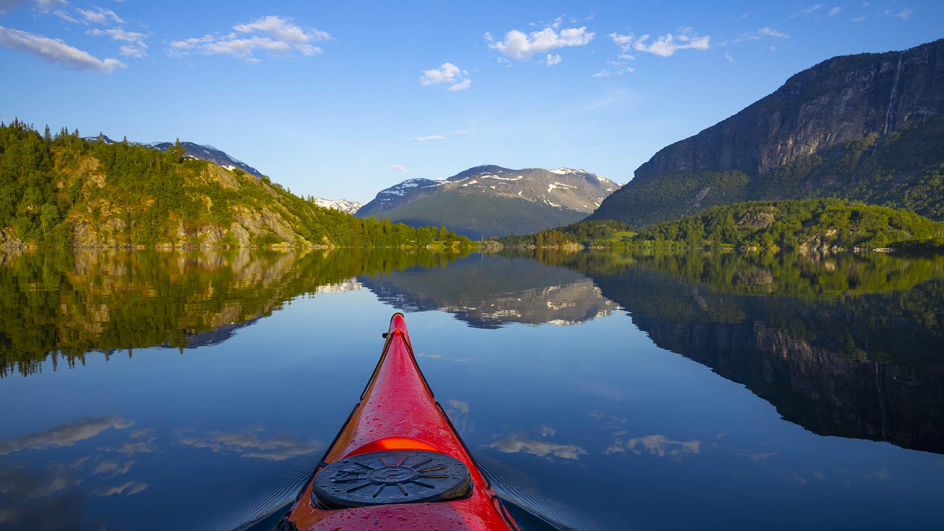 Paddeltour auf einem spiegelblanken See an einem herrlichen Sommermorgen. Die Ufer zu den Seiten sind grün, und im Hintergrund liegt ein Berg.