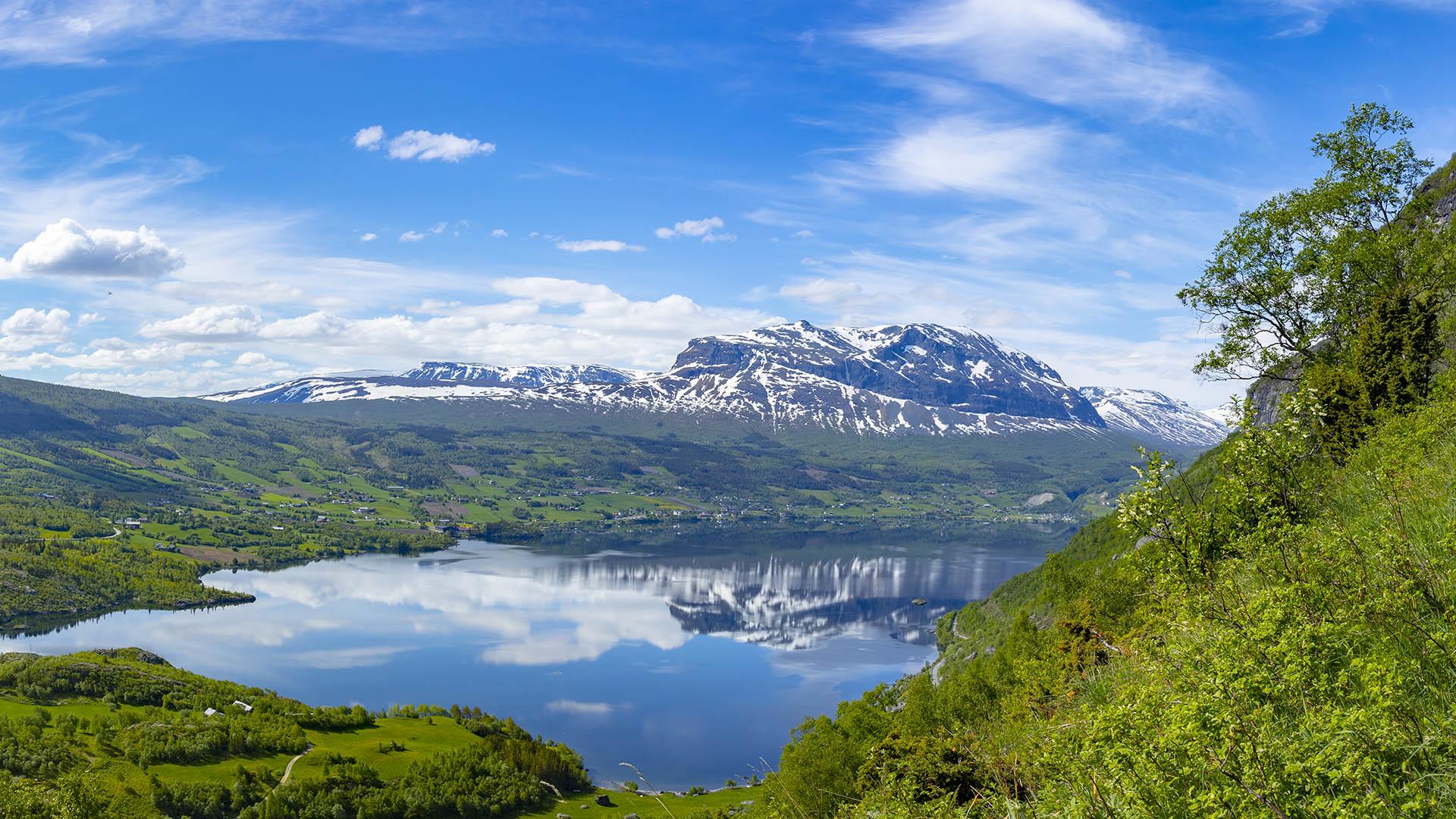 View from a green hill over a large blue lake. On the other side there are green fields and forets patches on the base of a large mountain massiv with still some snow. Blue sky and white summer clouds.