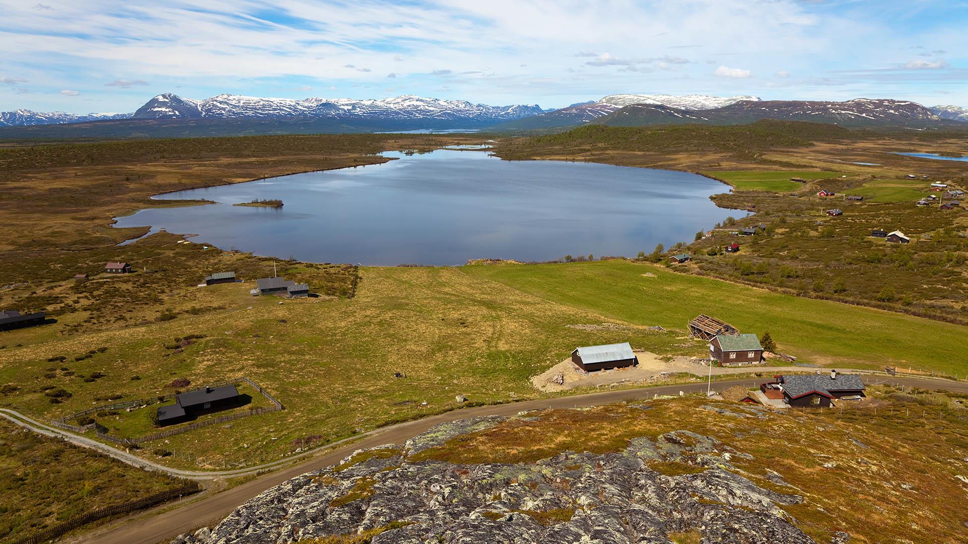 View from a hill over a high plateau above the tree line with a lake and some mountain farms. In the far horizon a mountain chain rises.