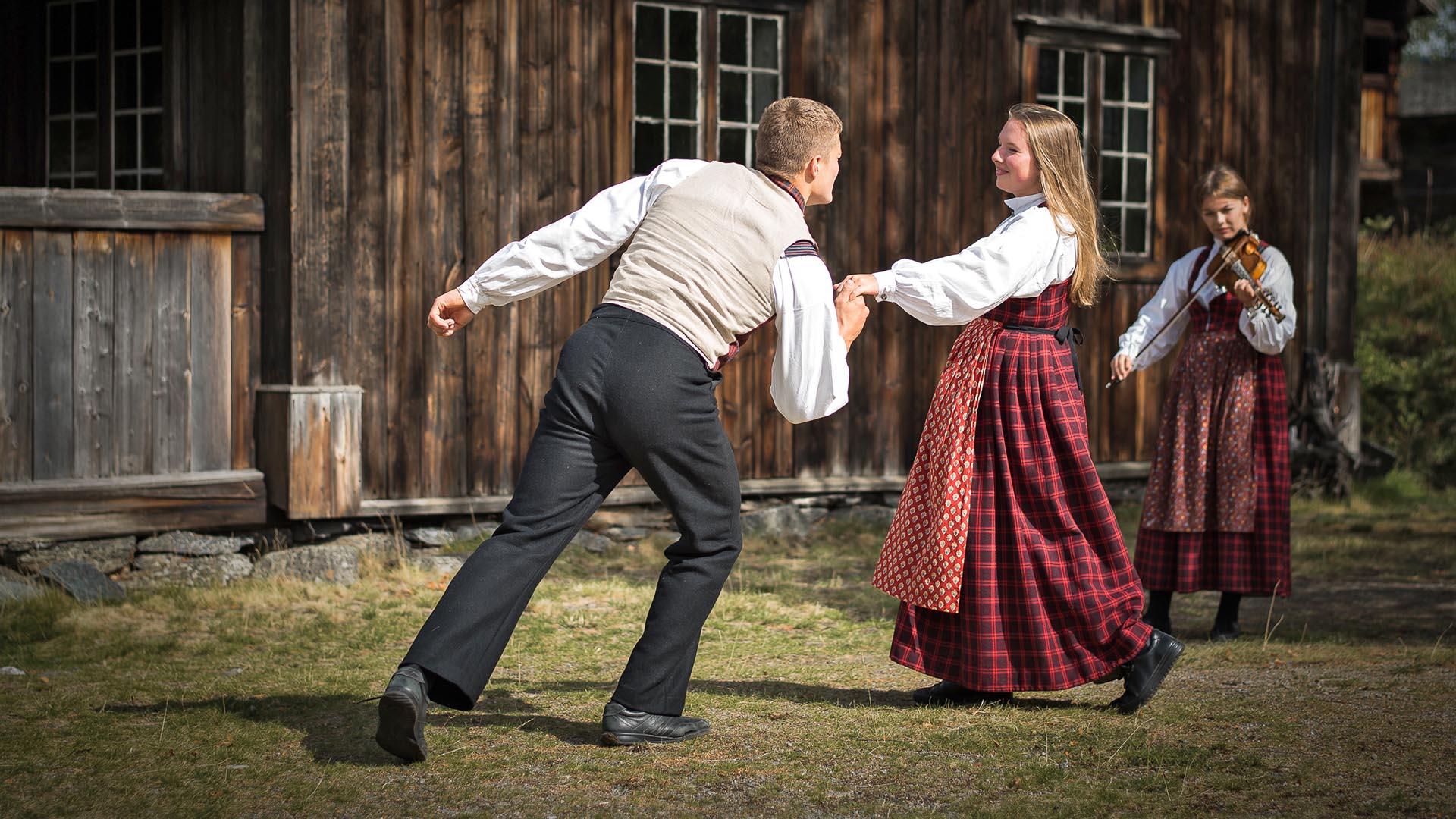 A young couple in folk costumes dances a folk dance on the lawn outside an old wooden building at Valdres Folk Museum, while another young girl plays the Hardanger fiddle.