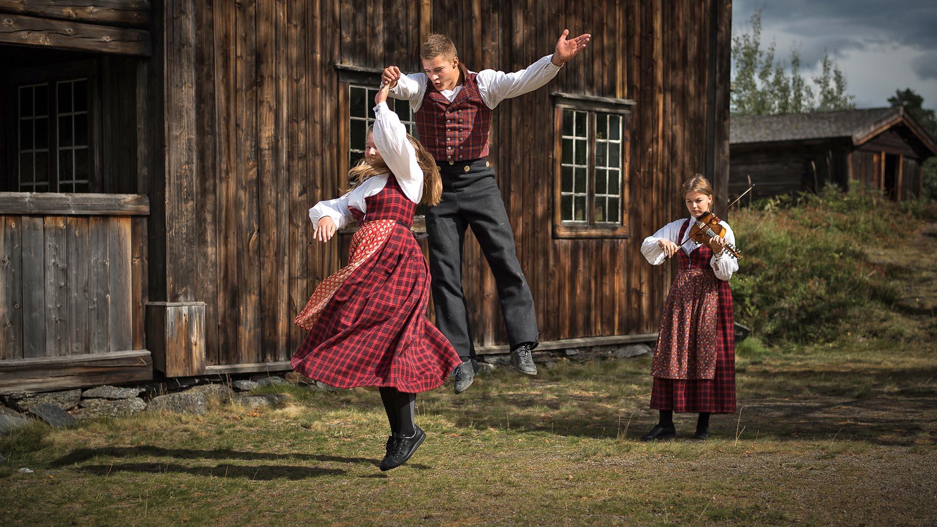 A young couple in folk costumes dances a folk dance on the lawn outside an old wooden building at Valdres Folk Museum. He jumps high, while she spins around. Another young girl plays the Hardanger fiddle.