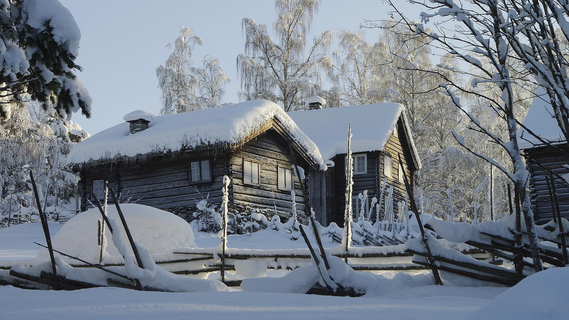 Two old log houses covered in snow behind a traditional roundpole fence.