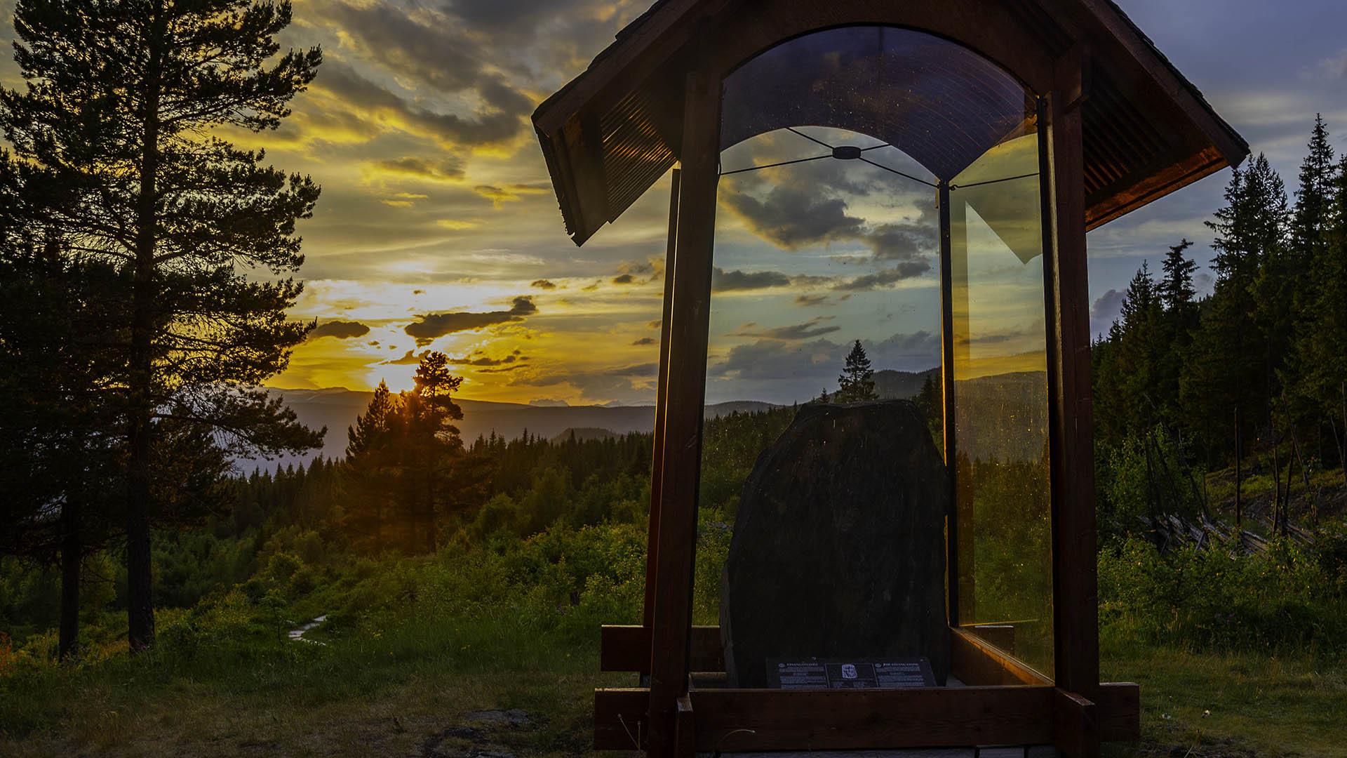 En runestein står i et glasshus på et gravfelt fra jernalderen. Sola går ned, og den oransje himmelen speiler seg i glasset.