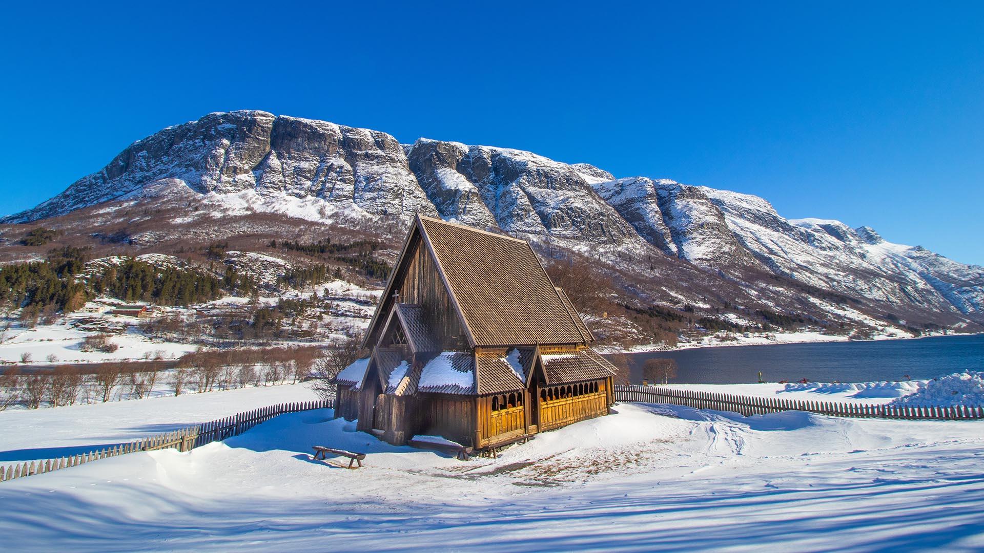 A small stave church on a snow-covered field with a lake and high mountains in the background. Blue sky.