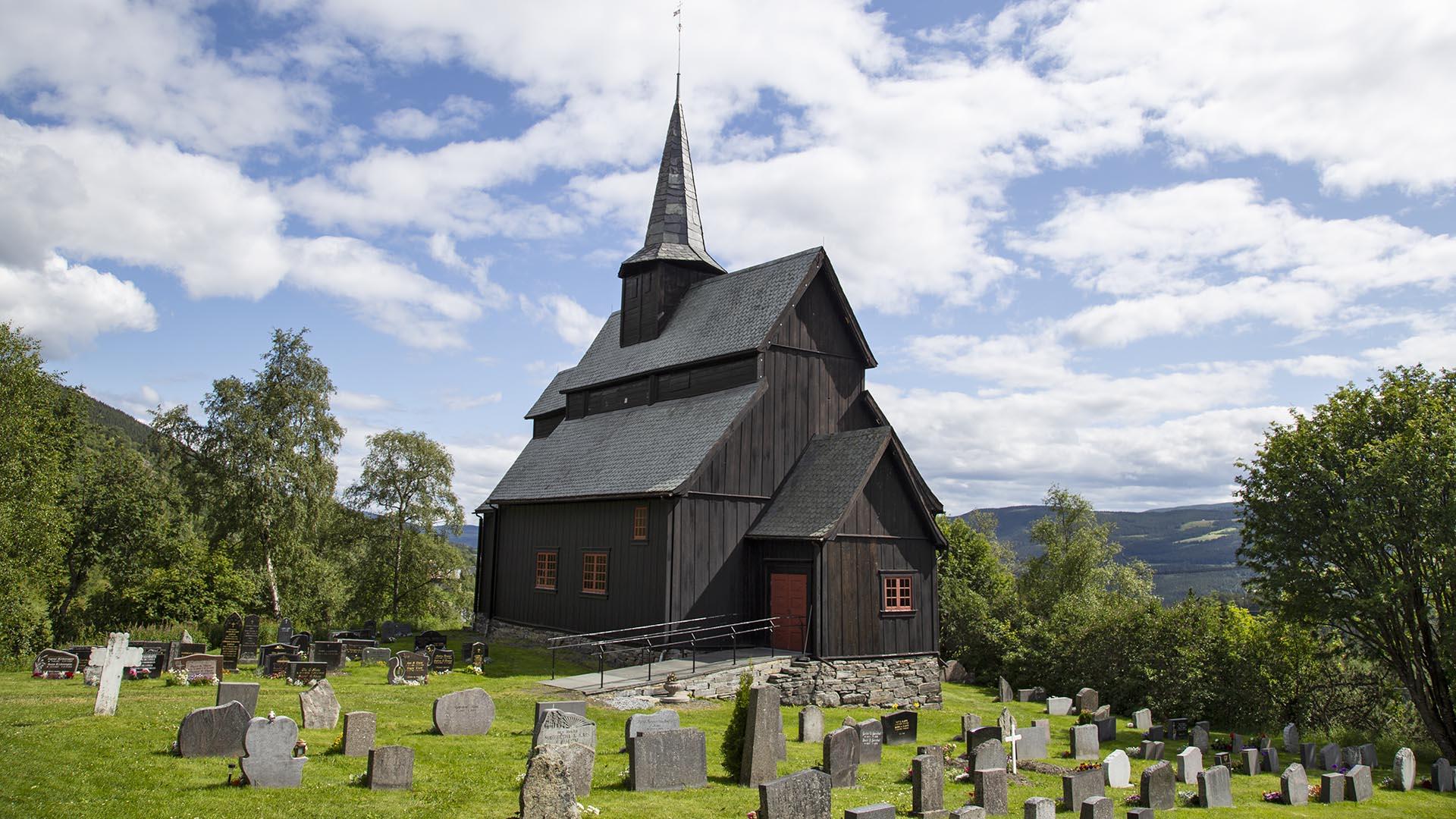 Gravstøtter i forgrunnen på en sommergrønn kirkegård med tjærebeiset stavkirke bak.