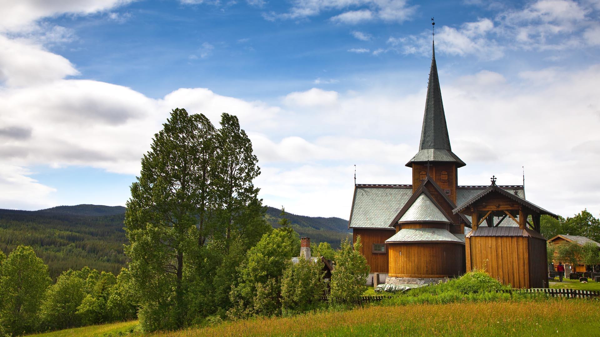 En sommerdag med grønn eng, grønne trær og en stavkirke.