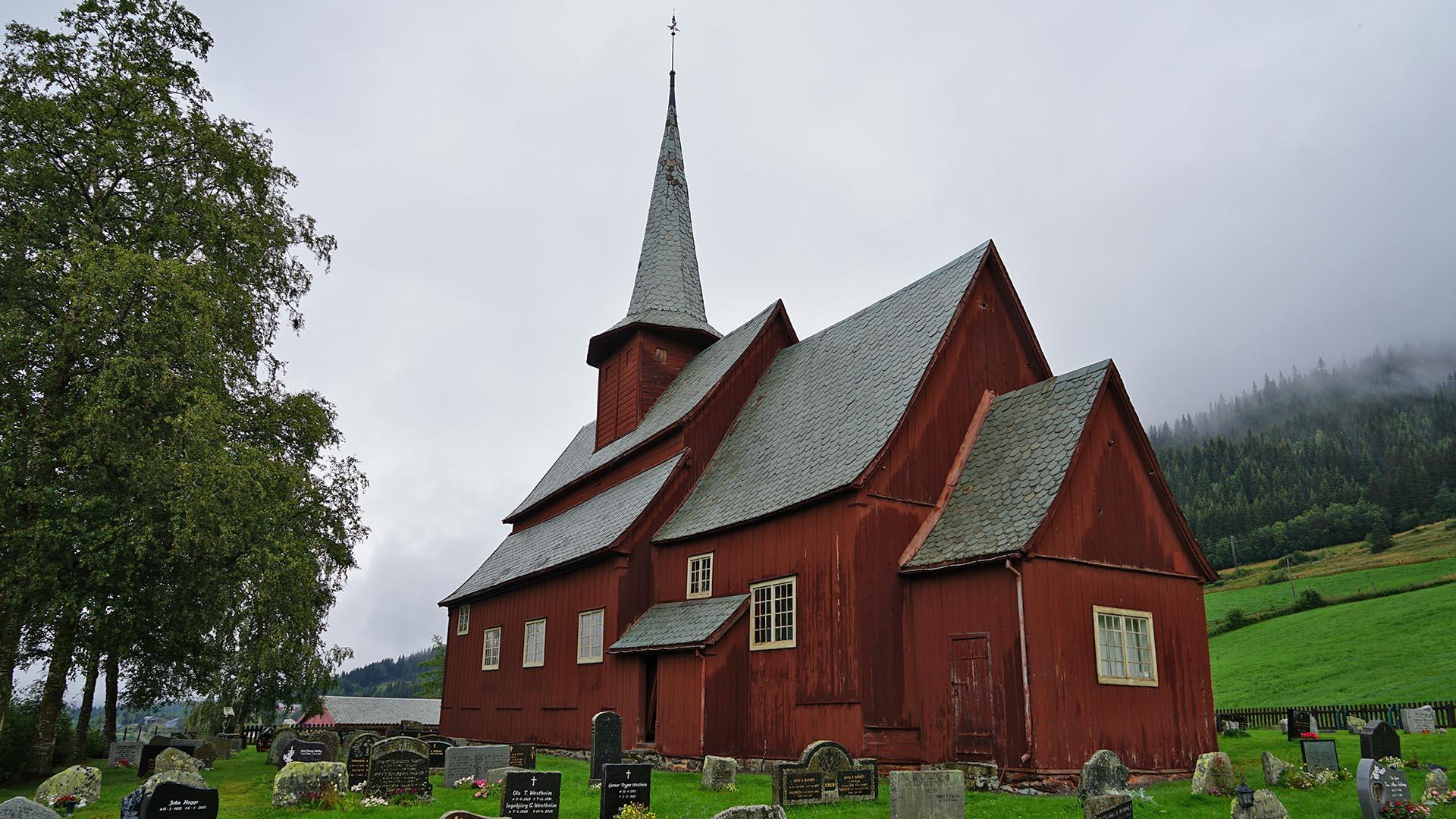 Gråværsdag på sommeren med grønt gress, en kirkegård og en liten burgundermalt stavkirke.