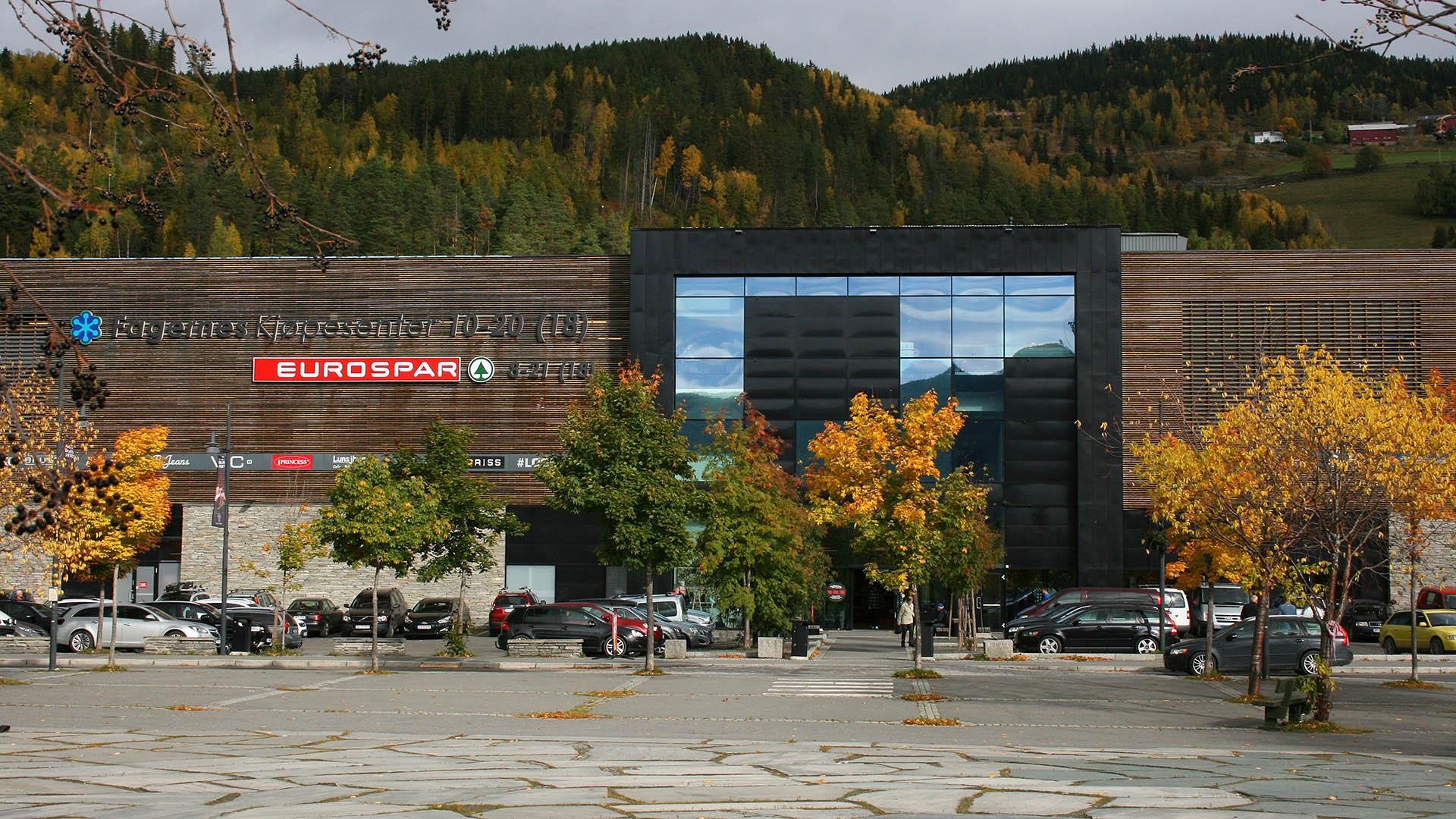 Trees on a plaza in front, a brown colored shopping mall in the background.